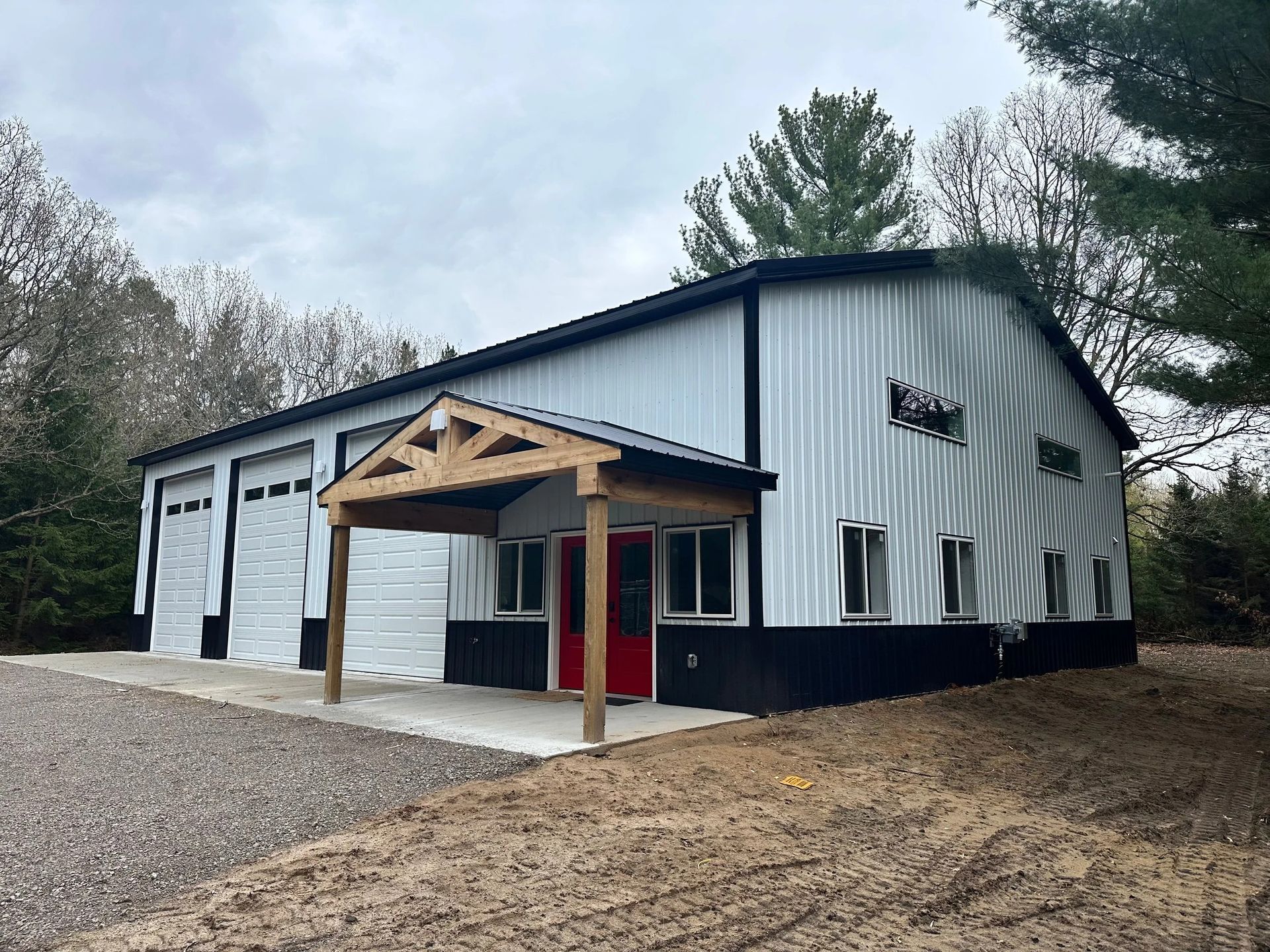 White metal workshop building with garage bays, red door, and covered entrance on a gravel lot