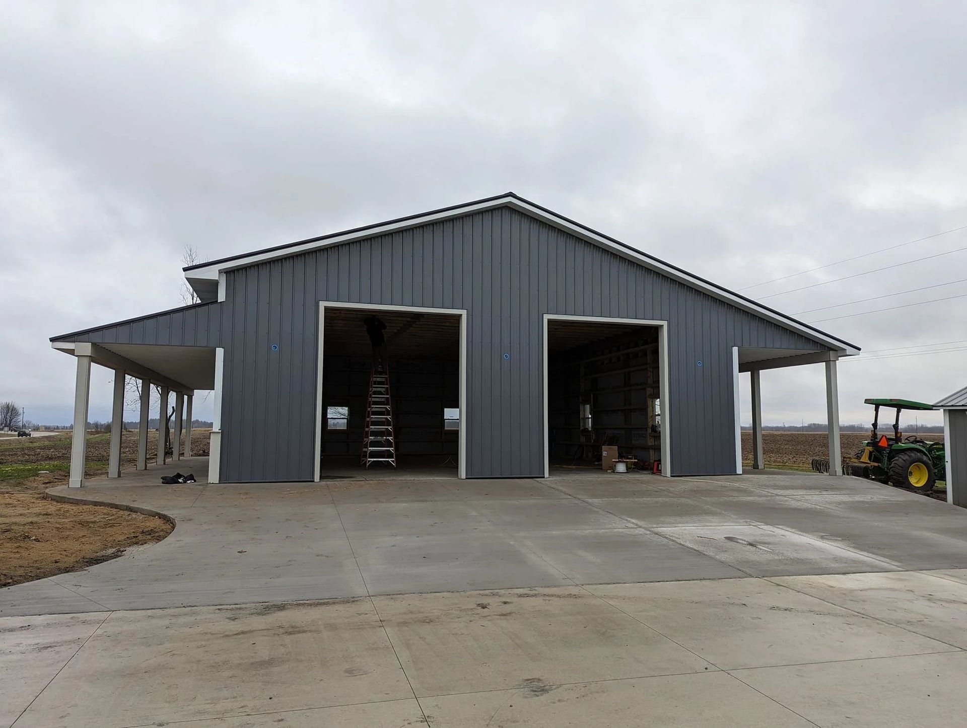 Gray metal barn with two open bays and a concrete driveway under an overcast sky