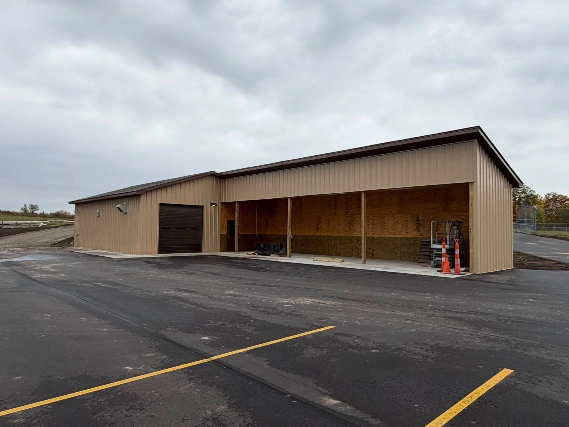 Empty tan metal building with an open bay on a paved lot under a cloudy sky