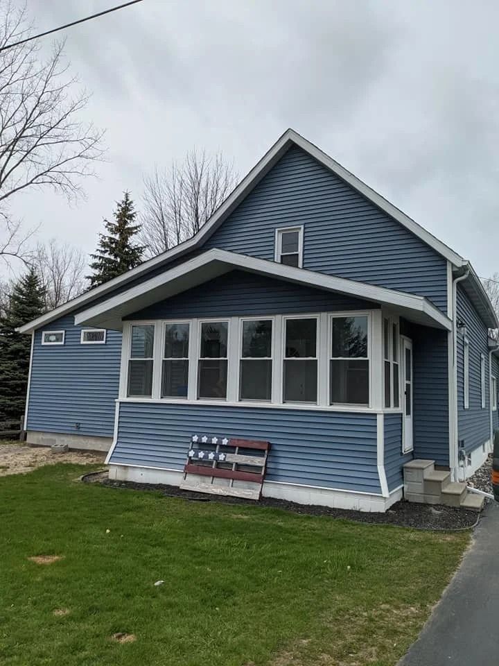Blue house with white trim, front porch windows, and a bench on a grassy lawn by a driveway
