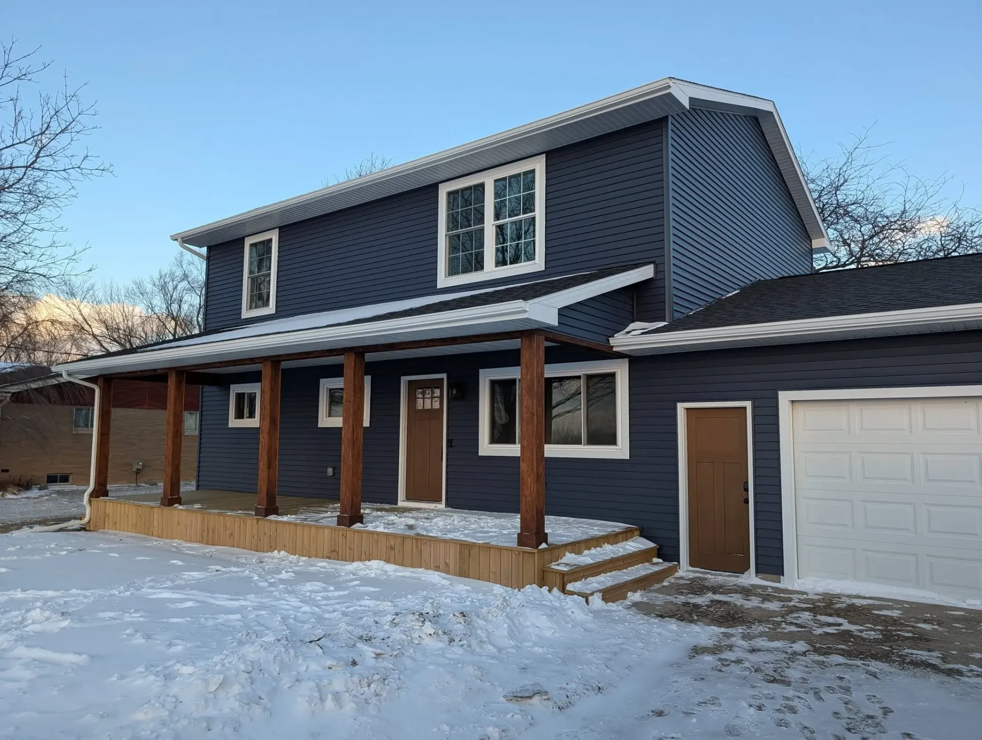 Snowy suburban house with dark blue siding, wood porch posts, and an attached garage