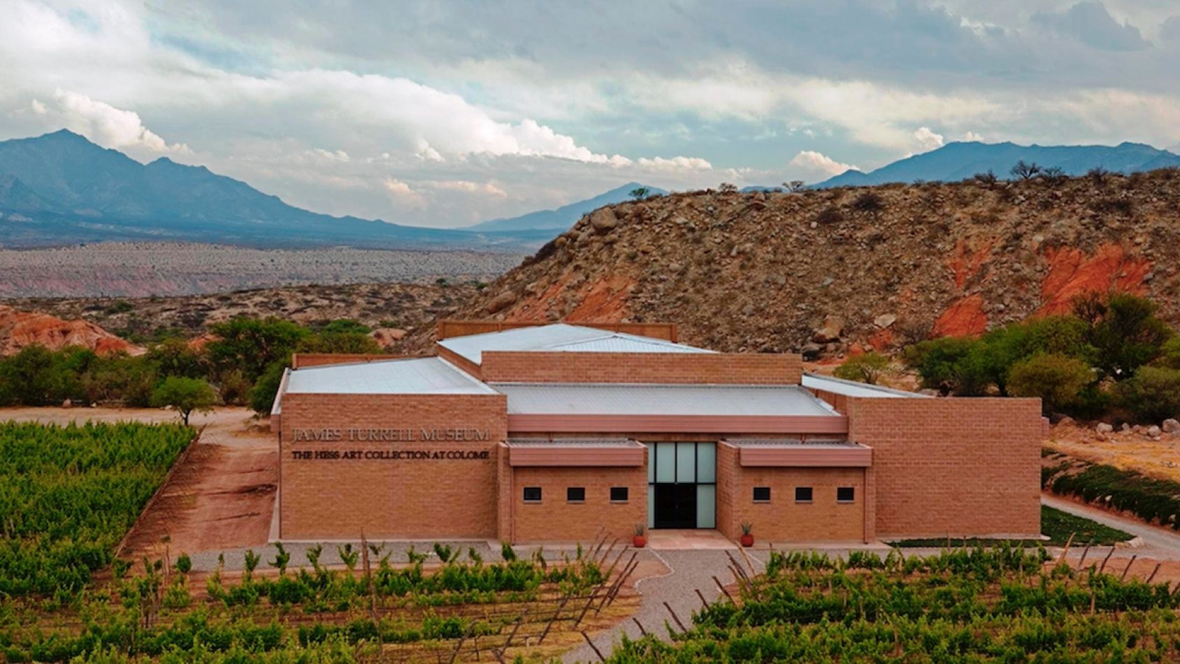 A large brick building is surrounded by a vineyard with mountains in the background.