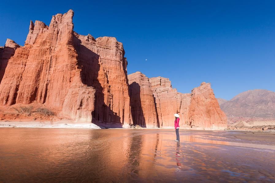 A woman in a red dress is standing on a beach in front of a large rock formation.