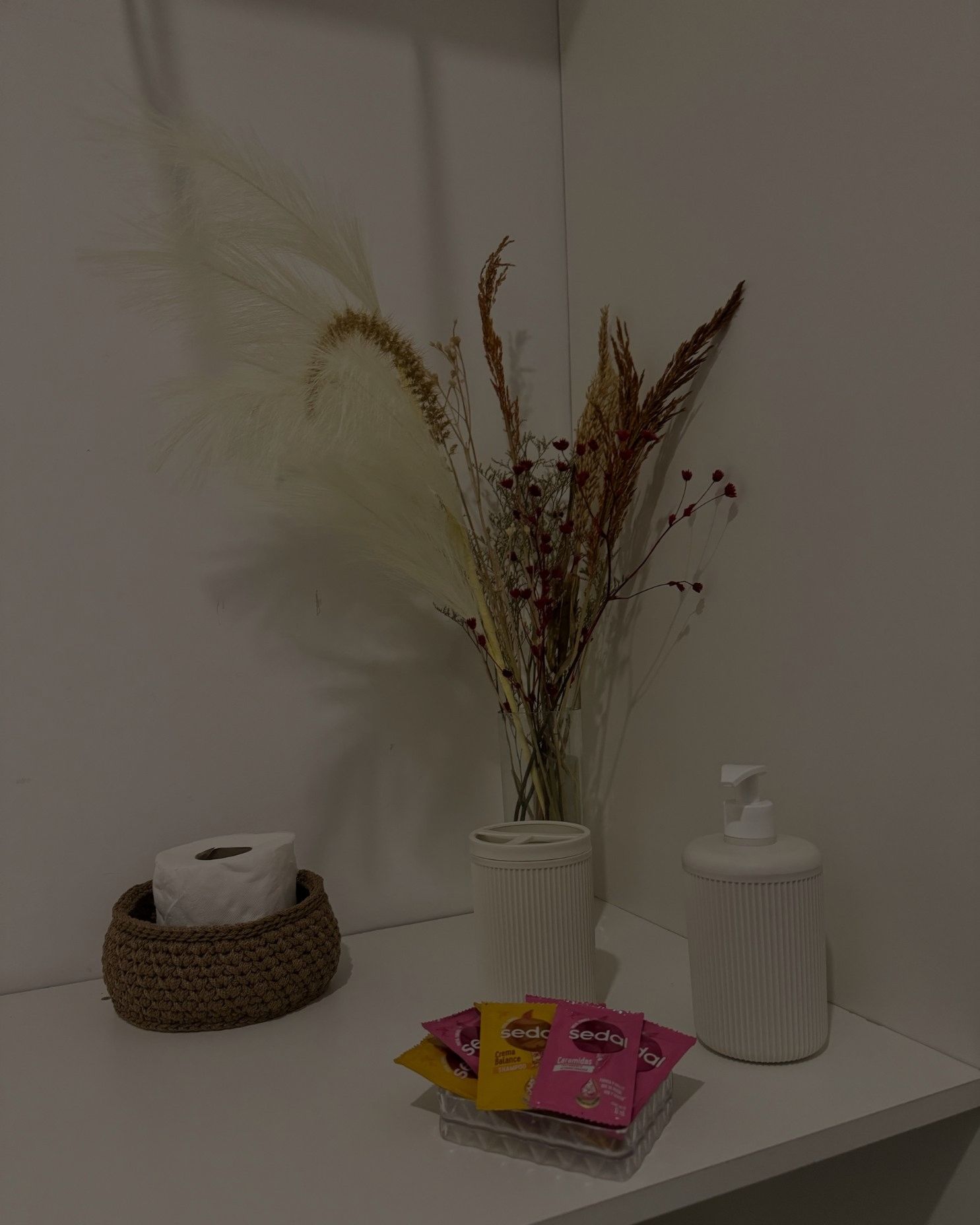 A bathroom shelf with a vase of flowers , tissues , and a soap dispenser.