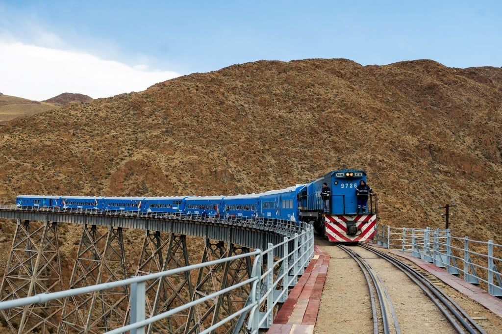 A blue train is going over a bridge in the desert.