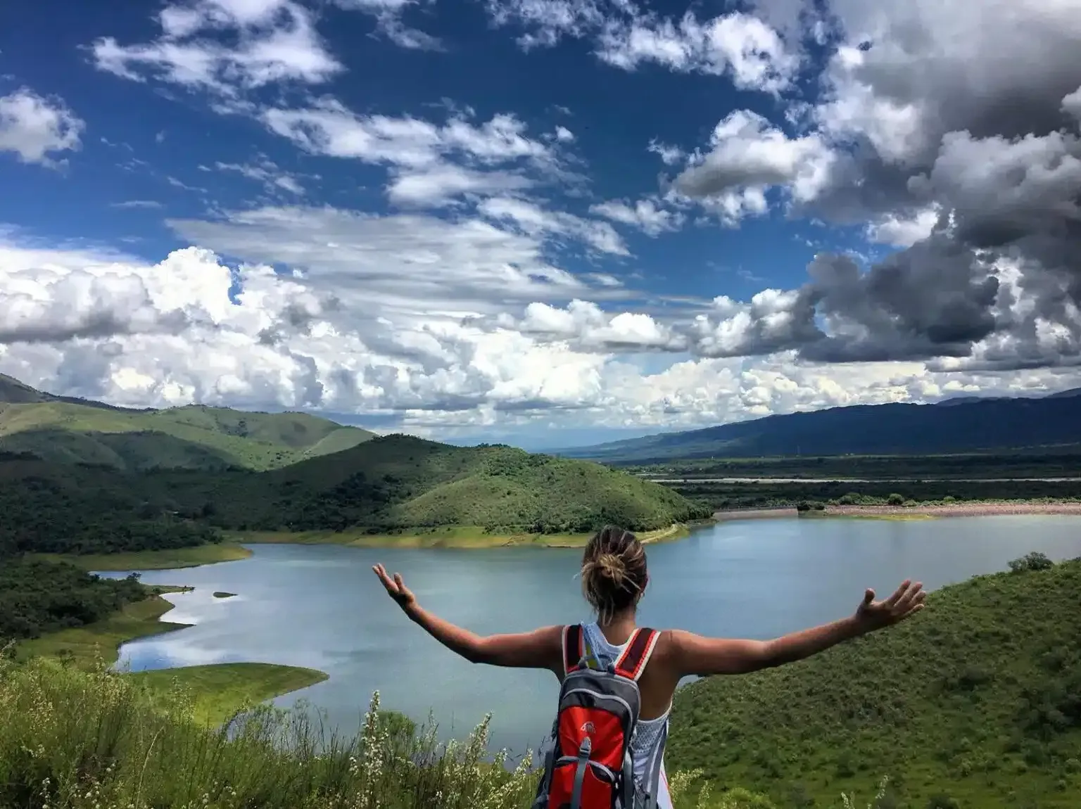 A woman with a backpack is standing on top of a hill overlooking a lake.