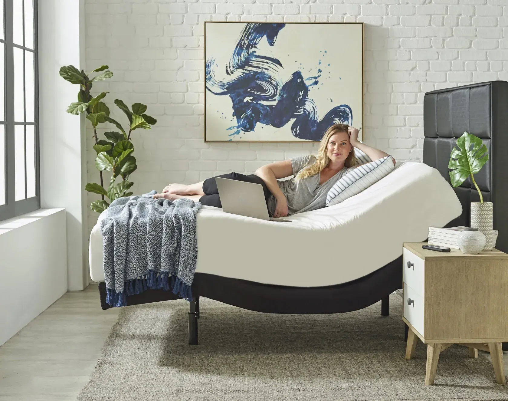 Woman lounging on an adjustable bed, using a laptop, in a bright bedroom.