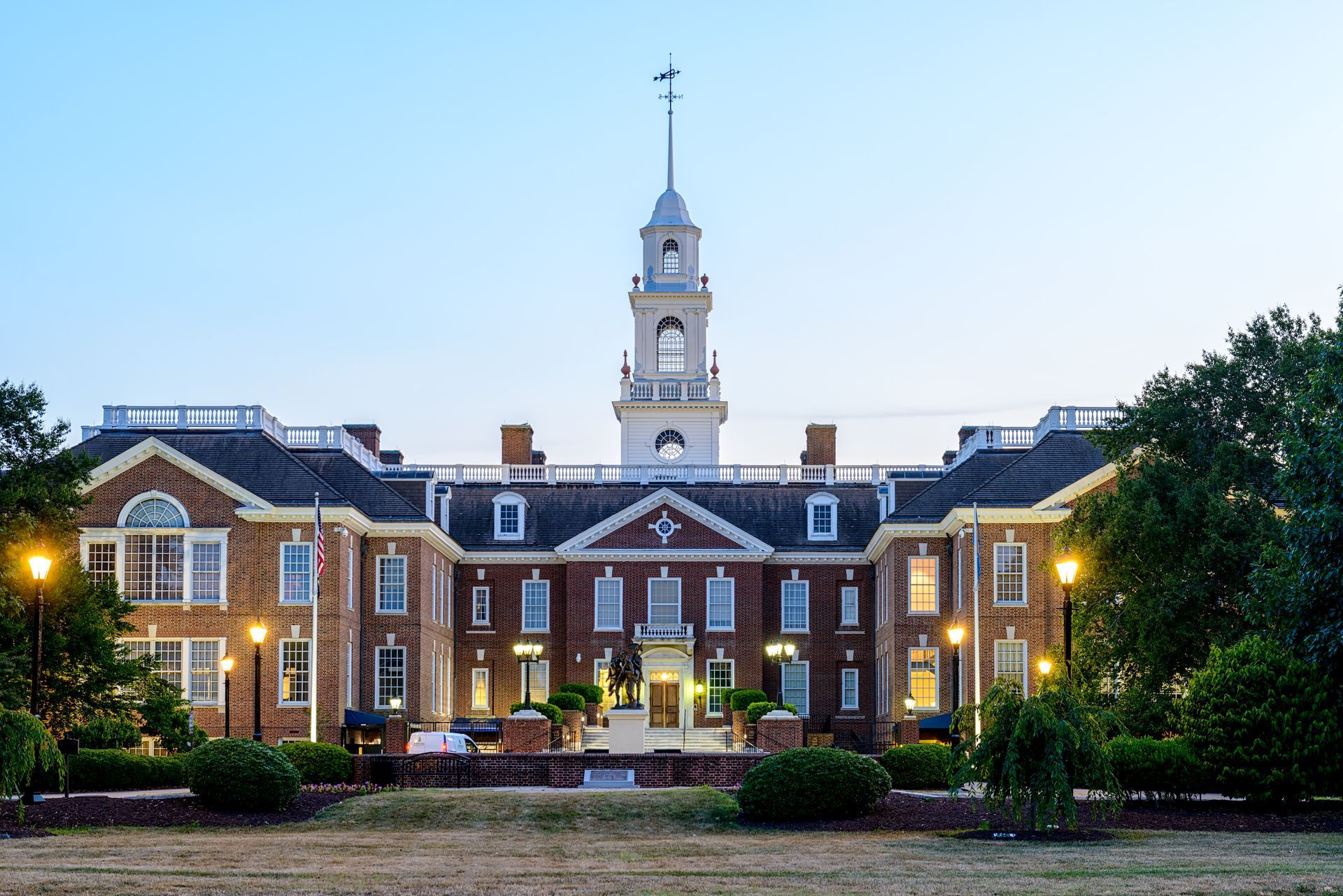 Historic brick building with central clock tower at dusk, framed by trees and lit windows