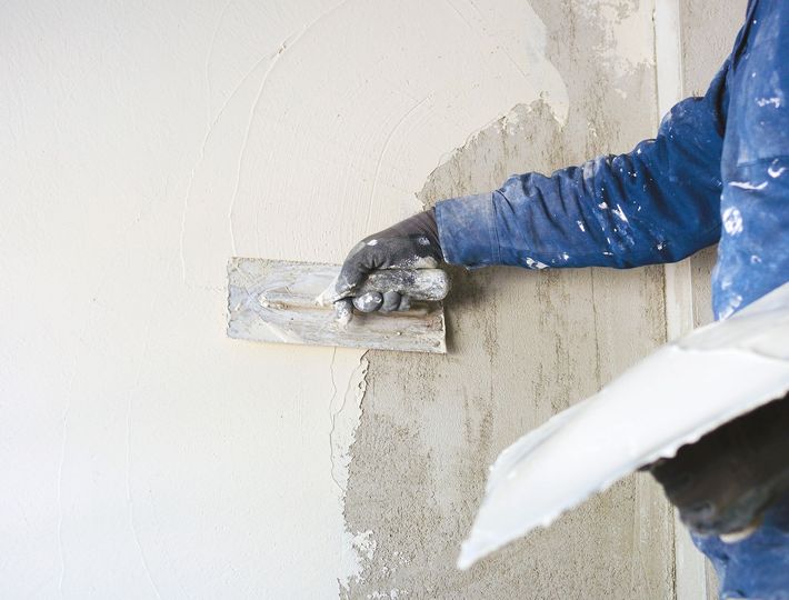 Worker applying plaster to a wall with a trowel, wearing a blue shirt and gloves.