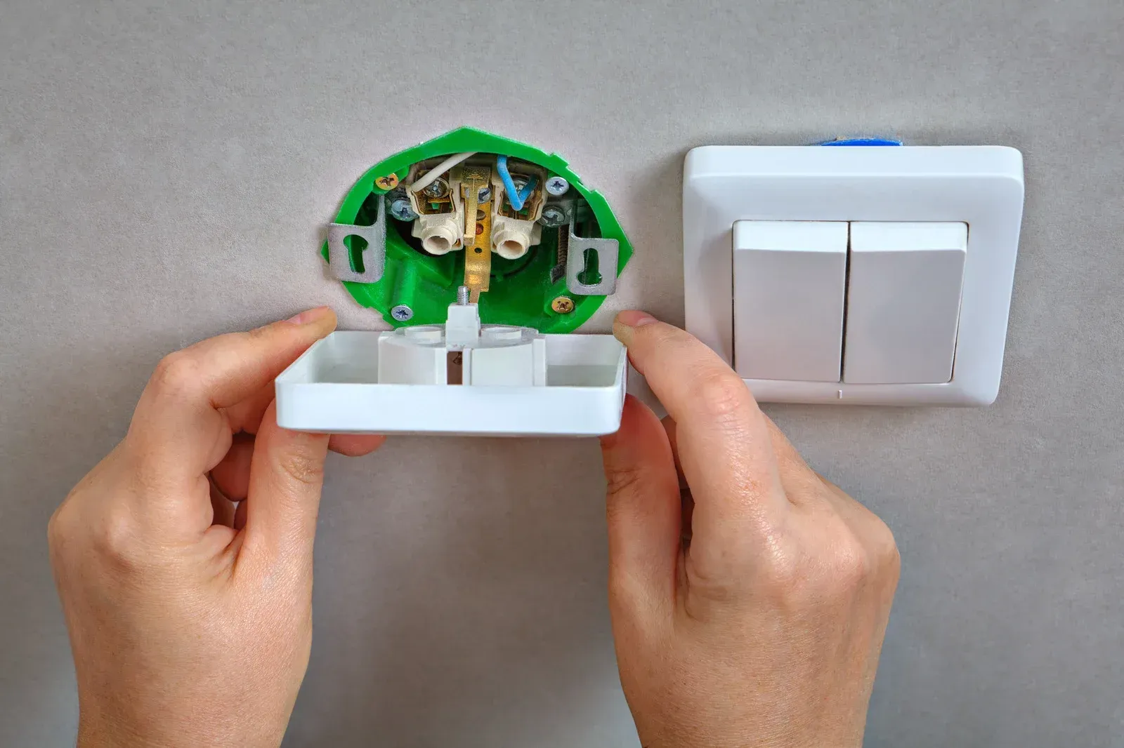 Hands installing a white light switch faceplate onto a green electrical box with exposed wires, next to a mounted switch.