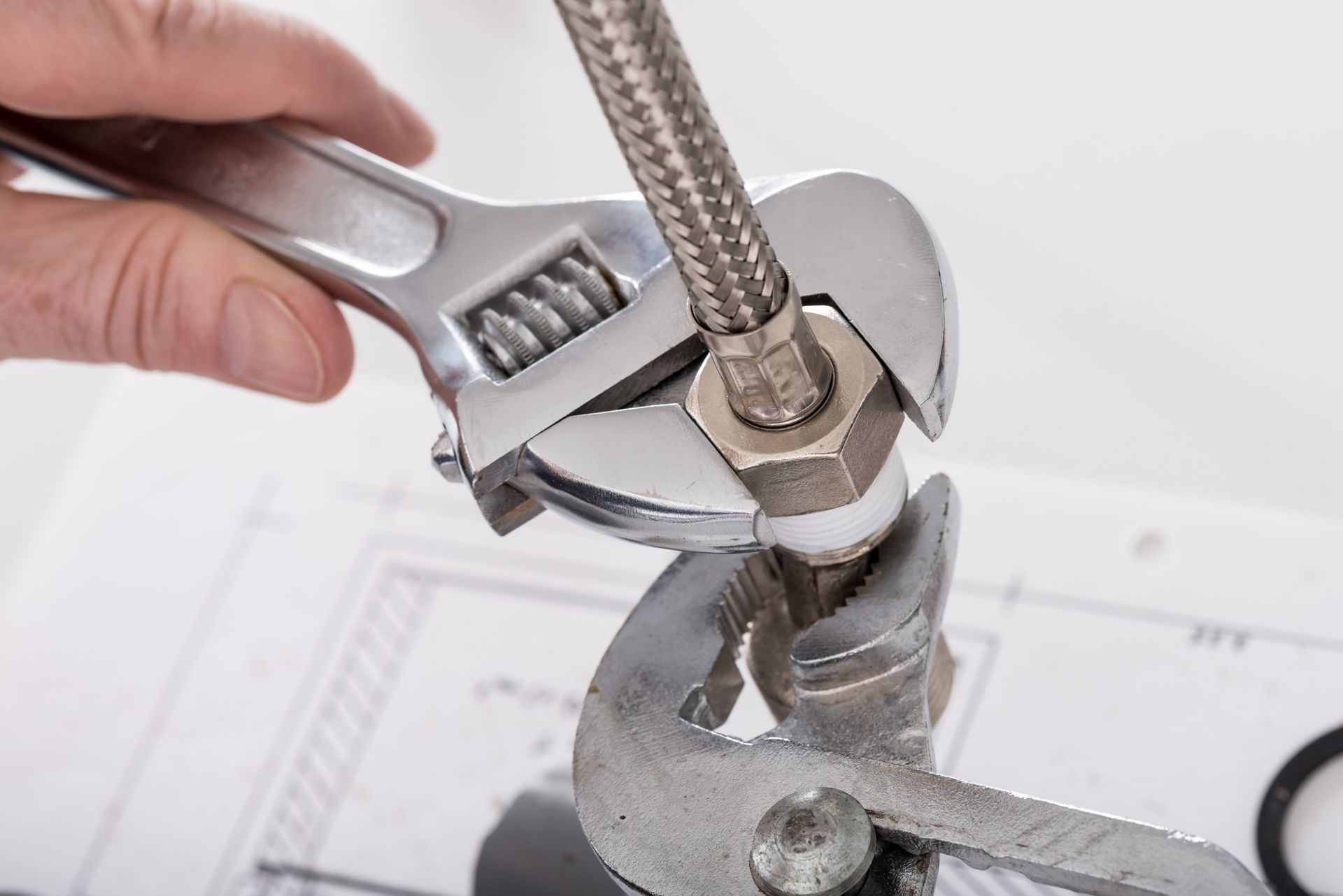 A hand uses an adjustable wrench to tighten a metal hose fitting held by pliers over technical blueprints.