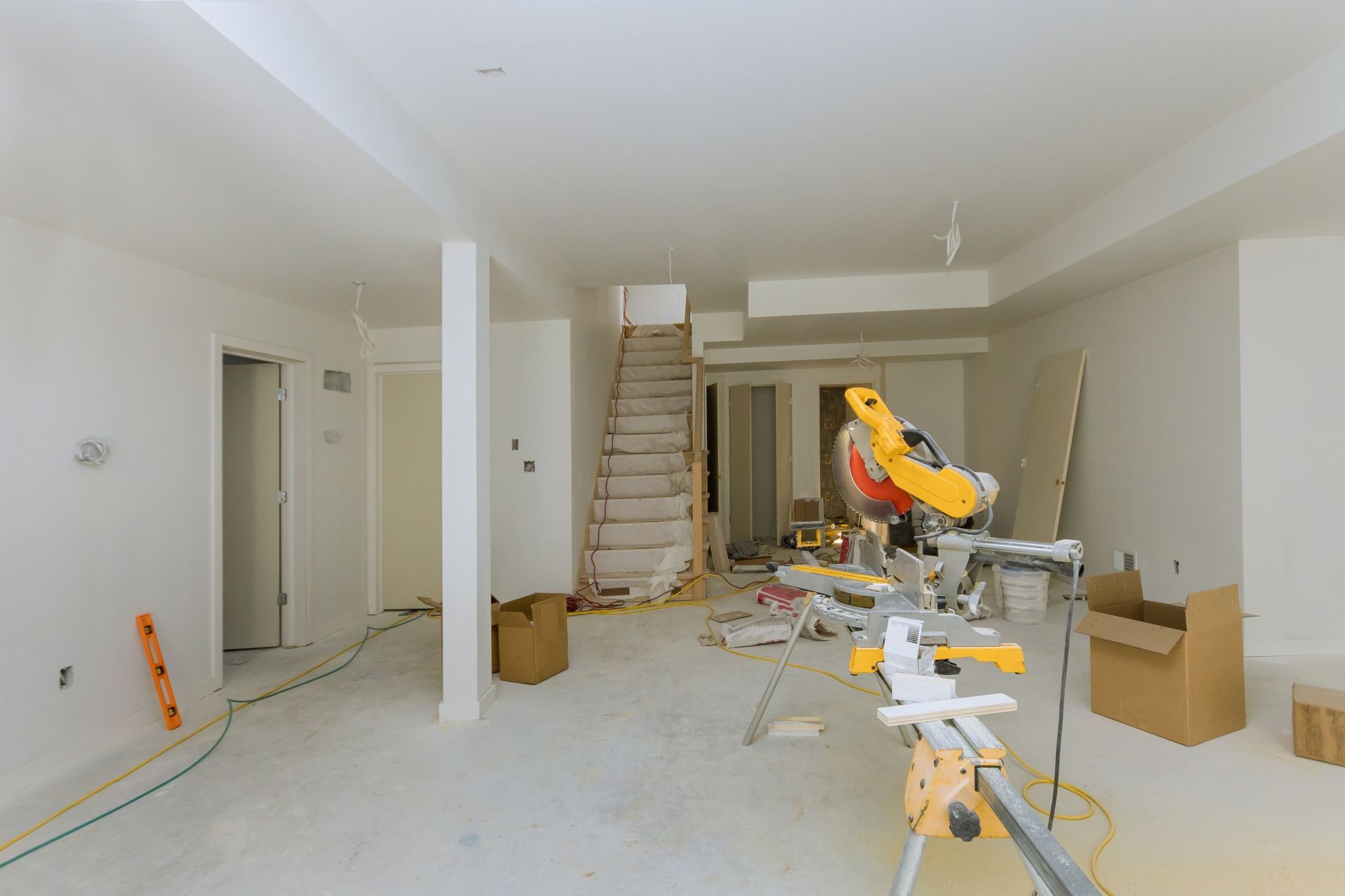 An unfinished interior room with white walls, a miter saw on a stand, boxes, and a staircase leading to an upper level.