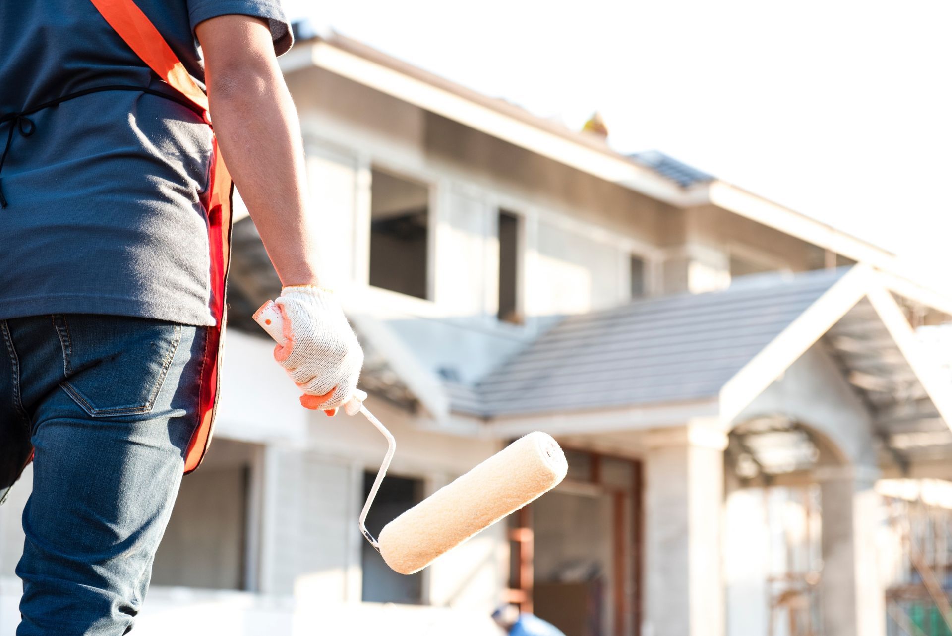 A person wearing a tool apron and holding a paint roller stands in front of a house under construction.
