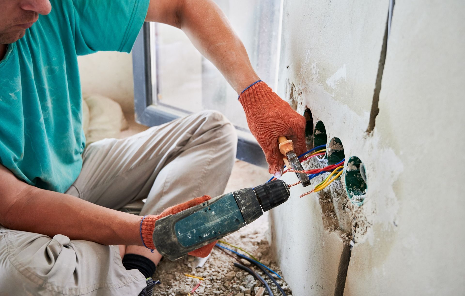 A worker wearing orange work gloves uses a drill to install electrical wiring into a wall box during home renovations.