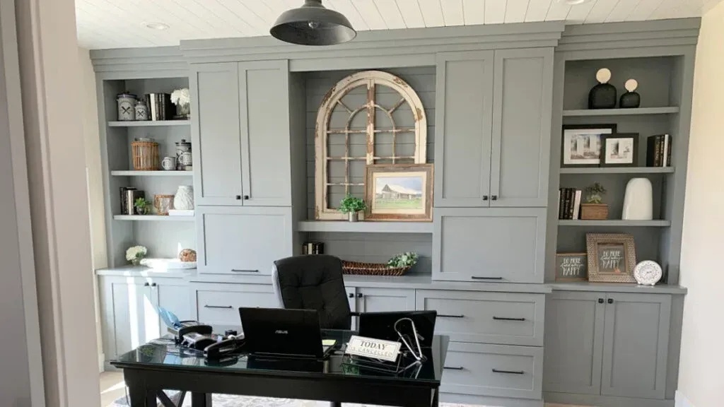 Built-in gray cabinetry with desk, open shelves, and arched window decor. Desk has a laptop and office supplies.
