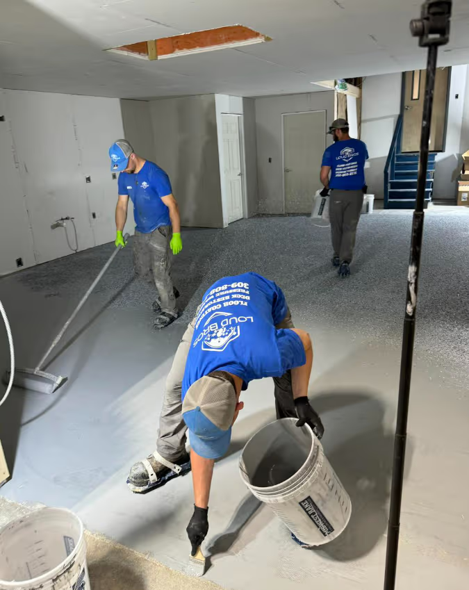 Three workers applying epoxy flooring in a concrete room. One pours from a bucket, the others spread the material.