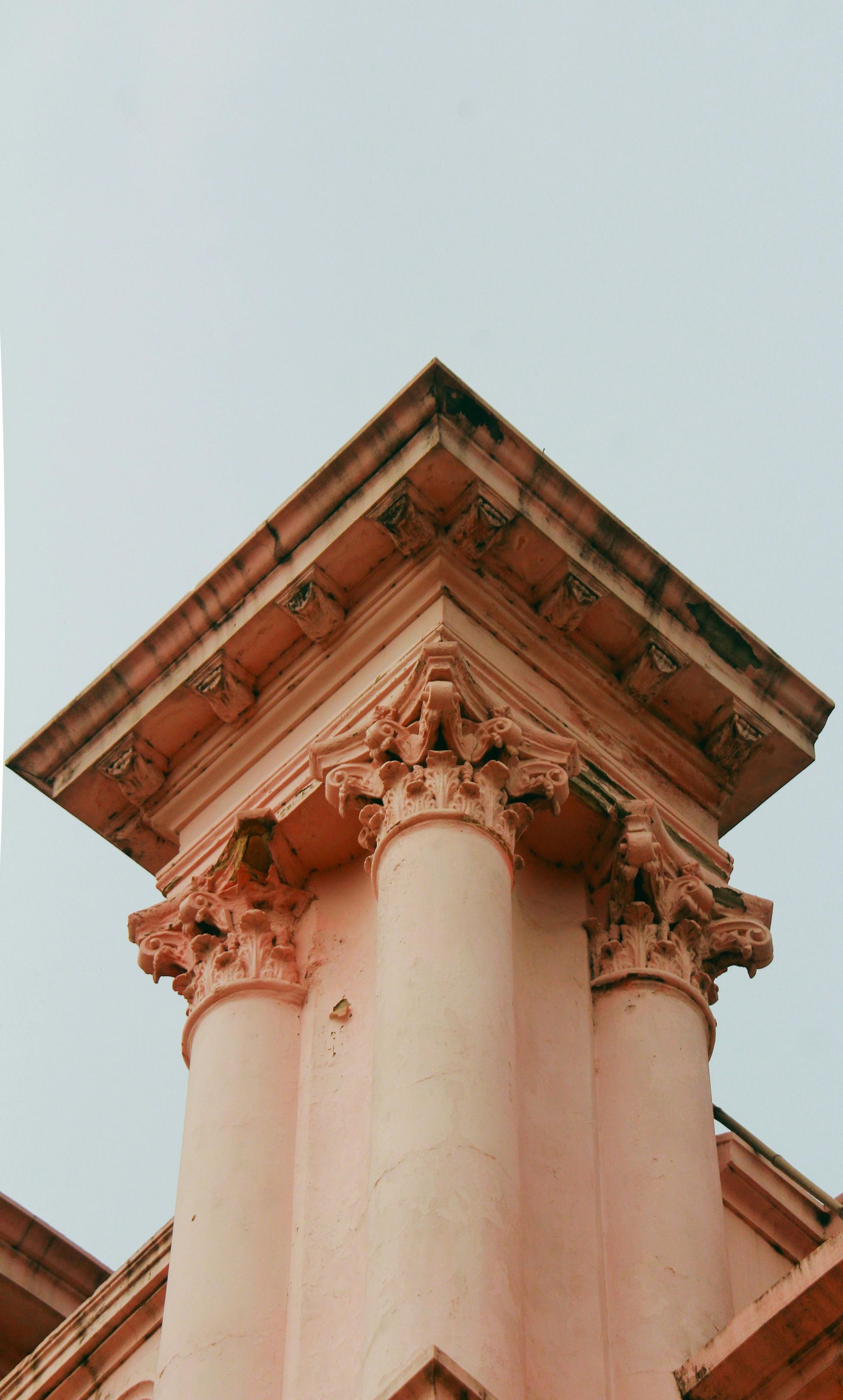 Upward view of a pink classical column with ornate capital and roofline against a pale sky