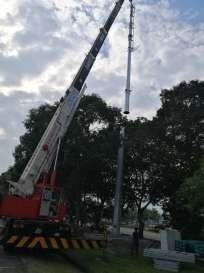 A crane lifting a tall metal pole to be placed on a base, near trees and a road.