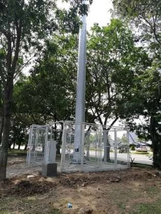 Tall silver cell tower in a park setting, surrounded by a fenced enclosure and trees.