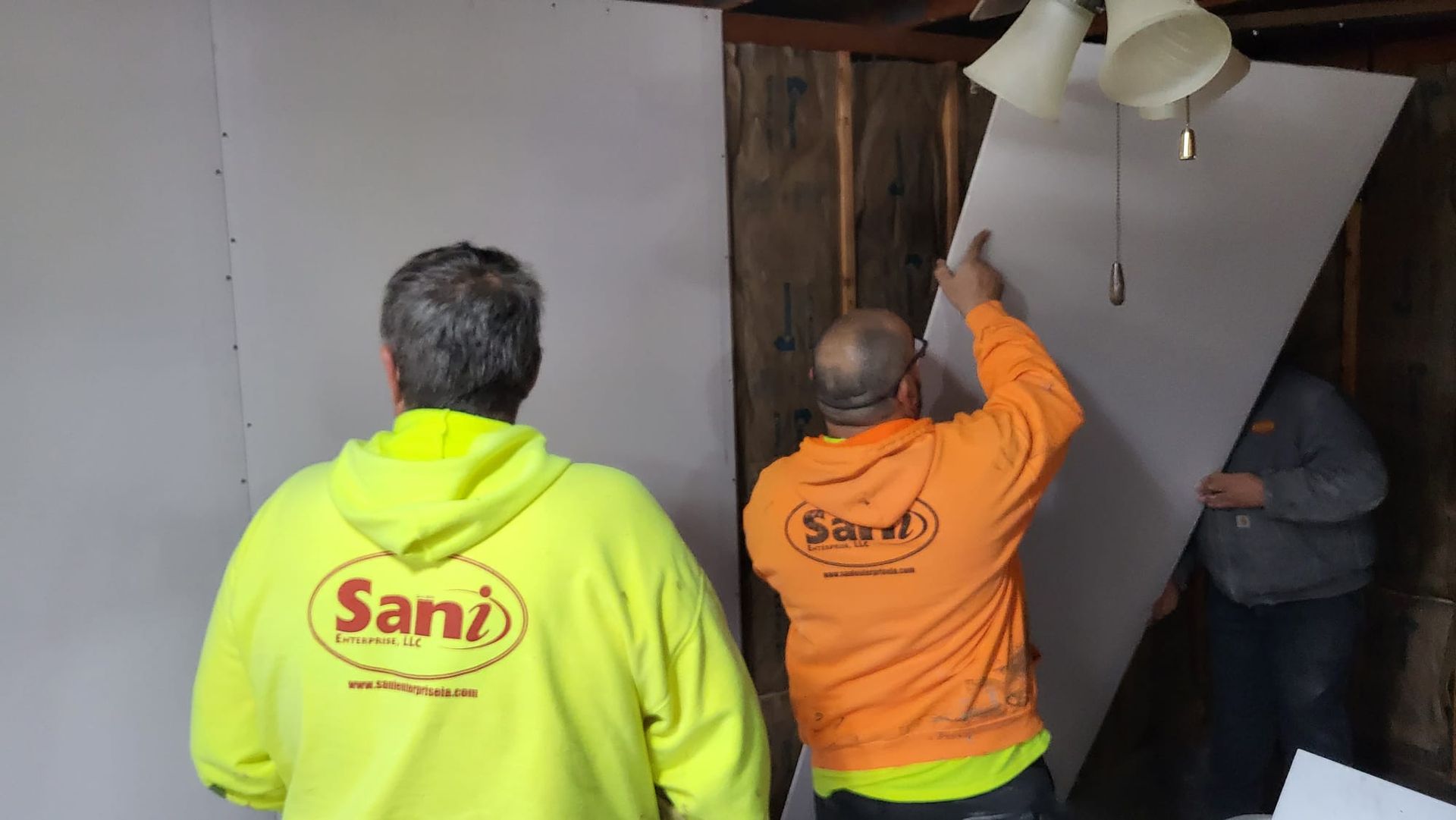 Two men are working on a wall and one of them is wearing an orange shirt that says sani.