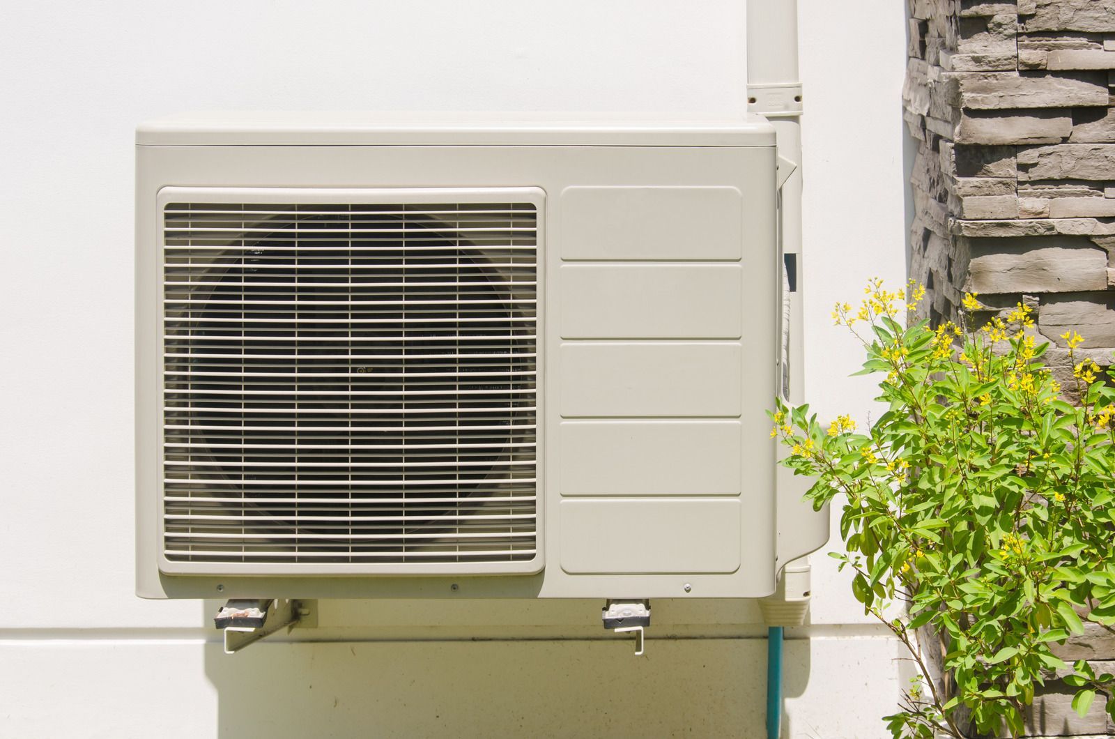 White air conditioning unit mounted on a white wall next to a brick column and green foliage.