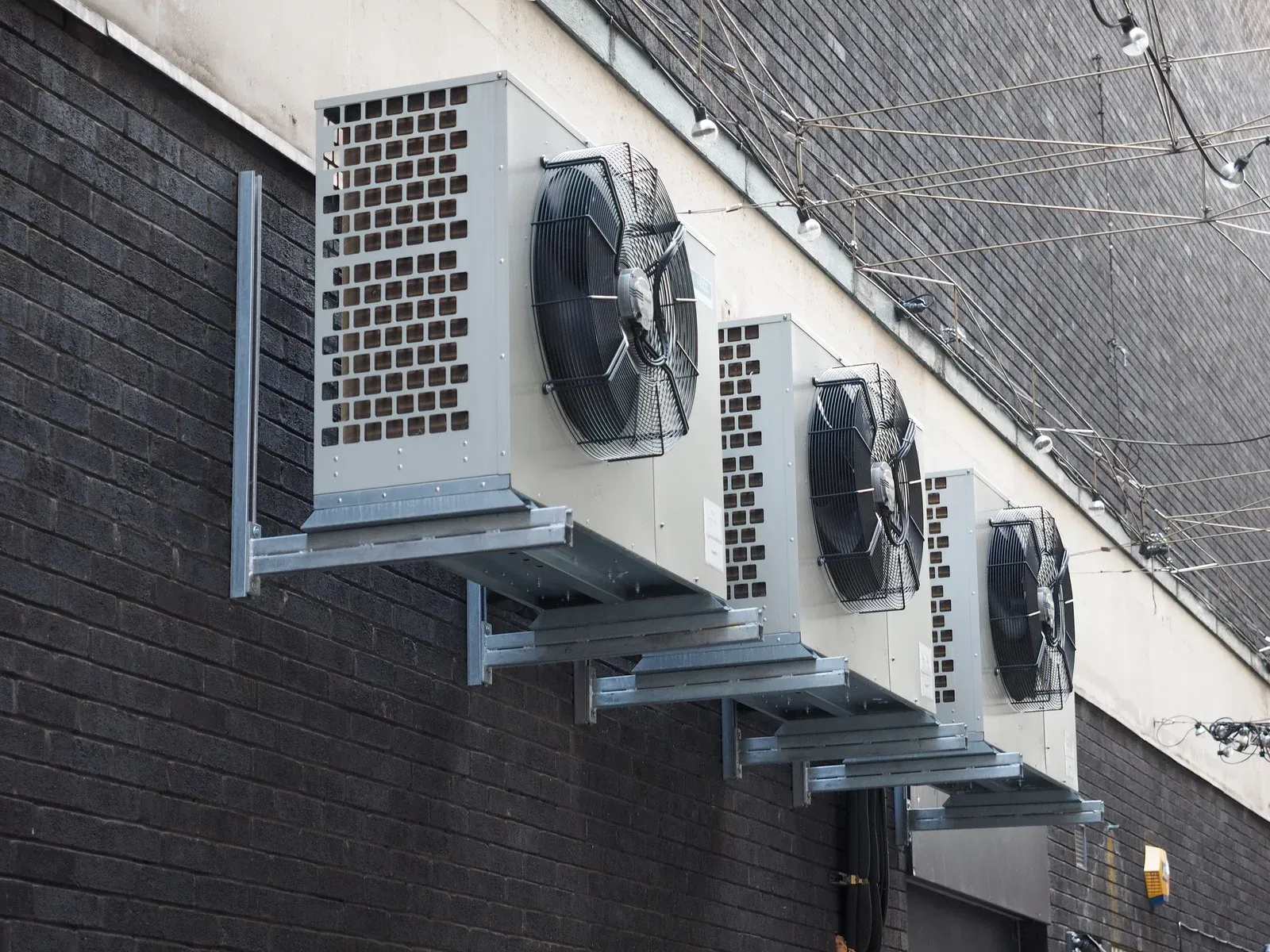 Four industrial air conditioning units mounted on a brick wall, with metal brackets.