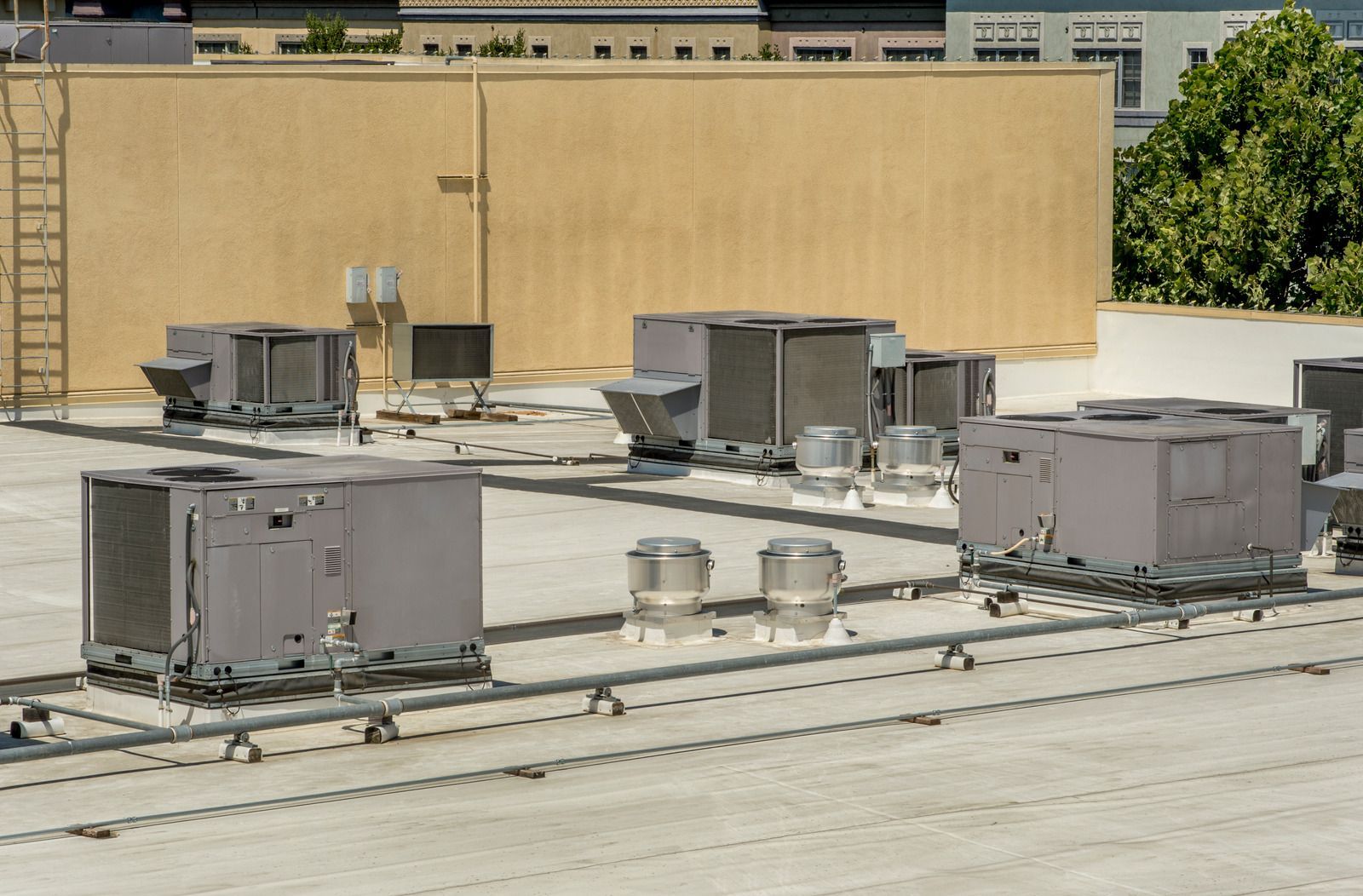 Air conditioning units on a flat rooftop, with vents and a ladder against a beige building.