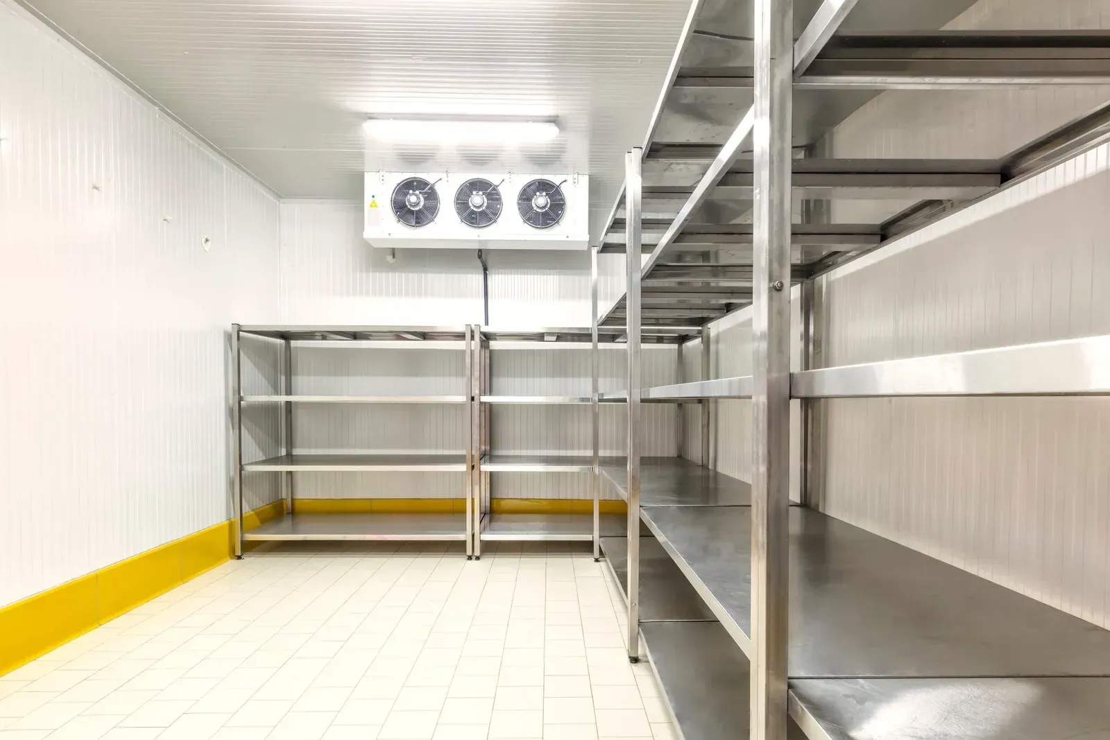 Empty walk-in freezer with stainless steel shelves, white walls, and a cooling unit.