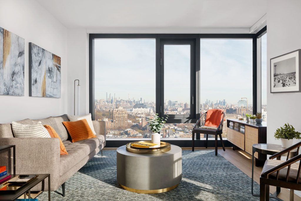 A bright living room with a gray sofa, a circular coffee table, and large windows offering a city skyline view.