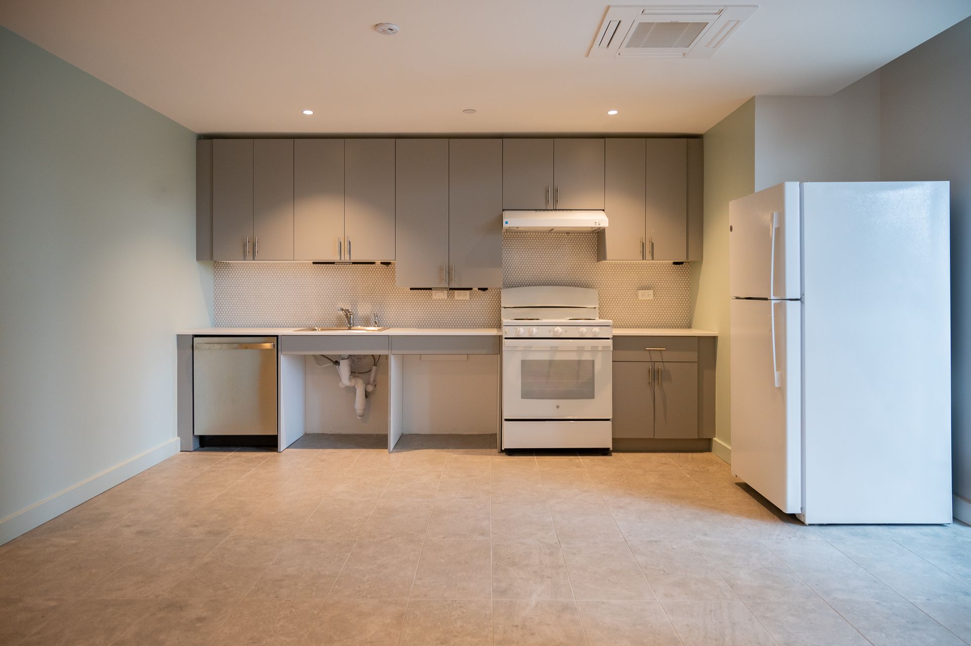 A modern, accessible kitchen with grey cabinets, white appliances, open under-sink space, and light beige tiled floors.