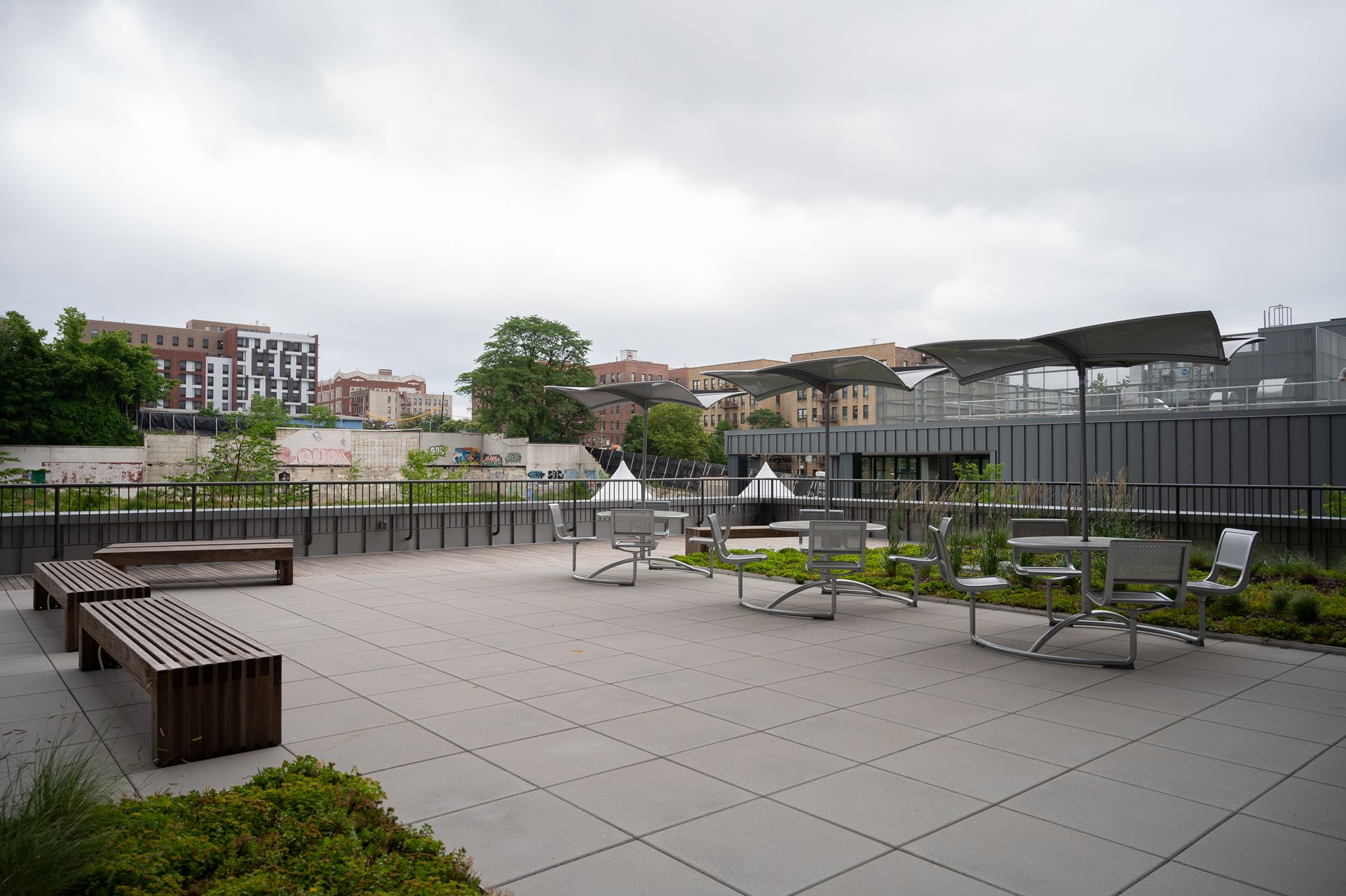An outdoor rooftop terrace featuring several metal benches, tables with umbrellas, and planters on a cloudy day.