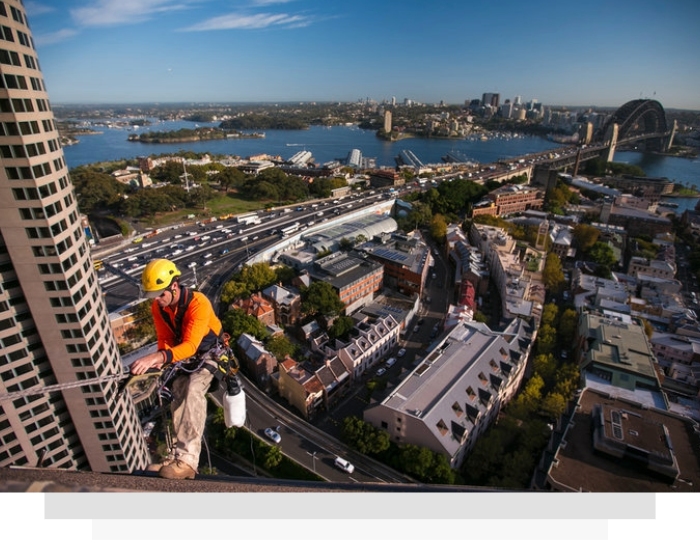 Roofer in orange safety vest on a high-rise ledge overlooking Sydney Harbour and the Harbour Bridge