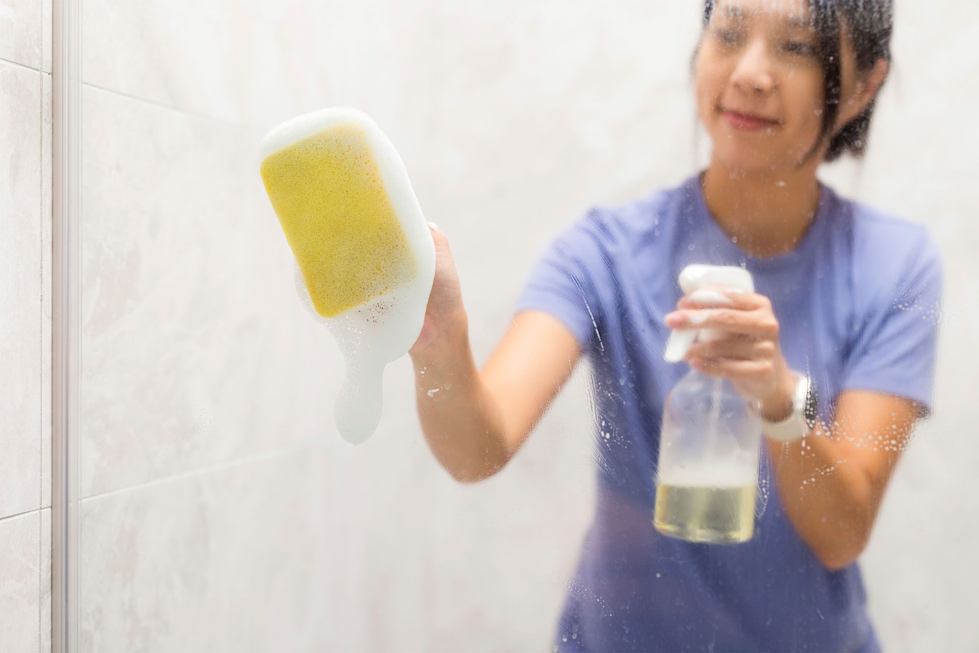 A woman is cleaning a shower door with a sponge and spray bottle.