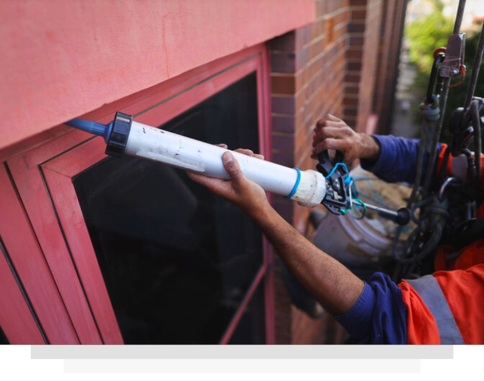 A worker suspended in a harness uses a caulk gun to apply sealant around the frame of a window on a brick building.