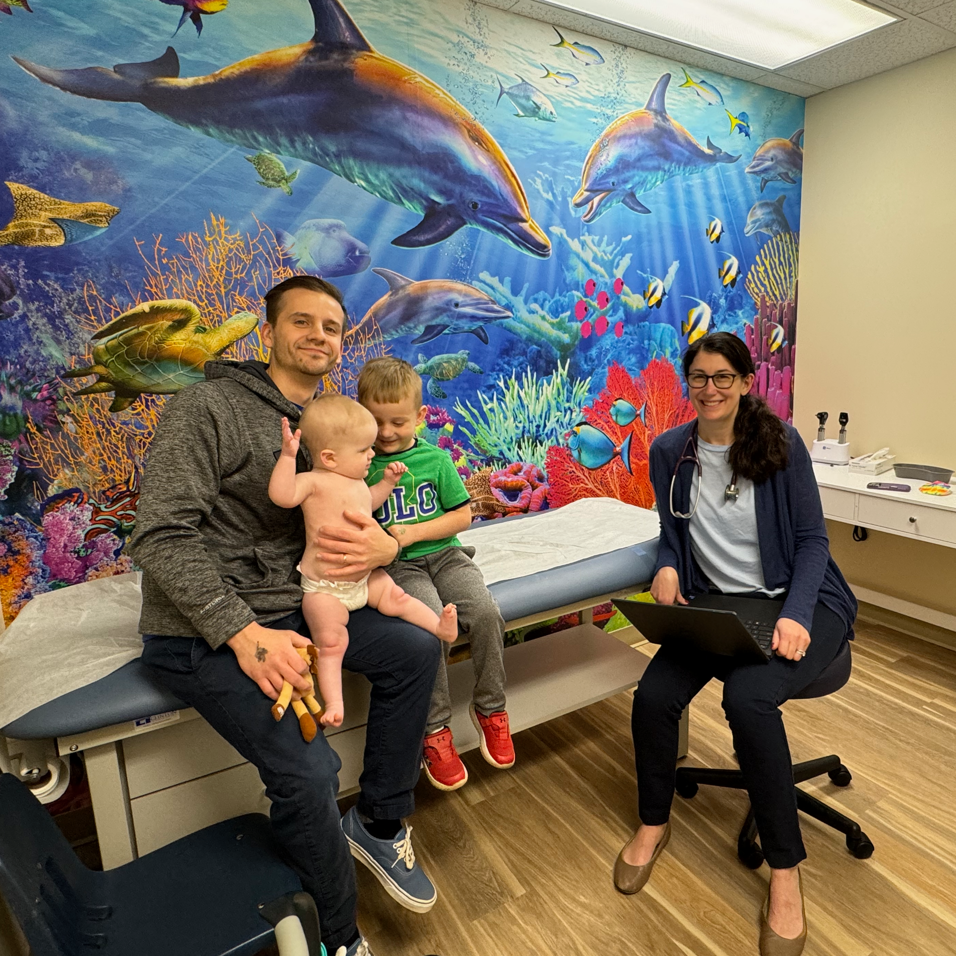 A man and a woman are sitting in a medical exam room with two children in front of a dolphin wall mural.