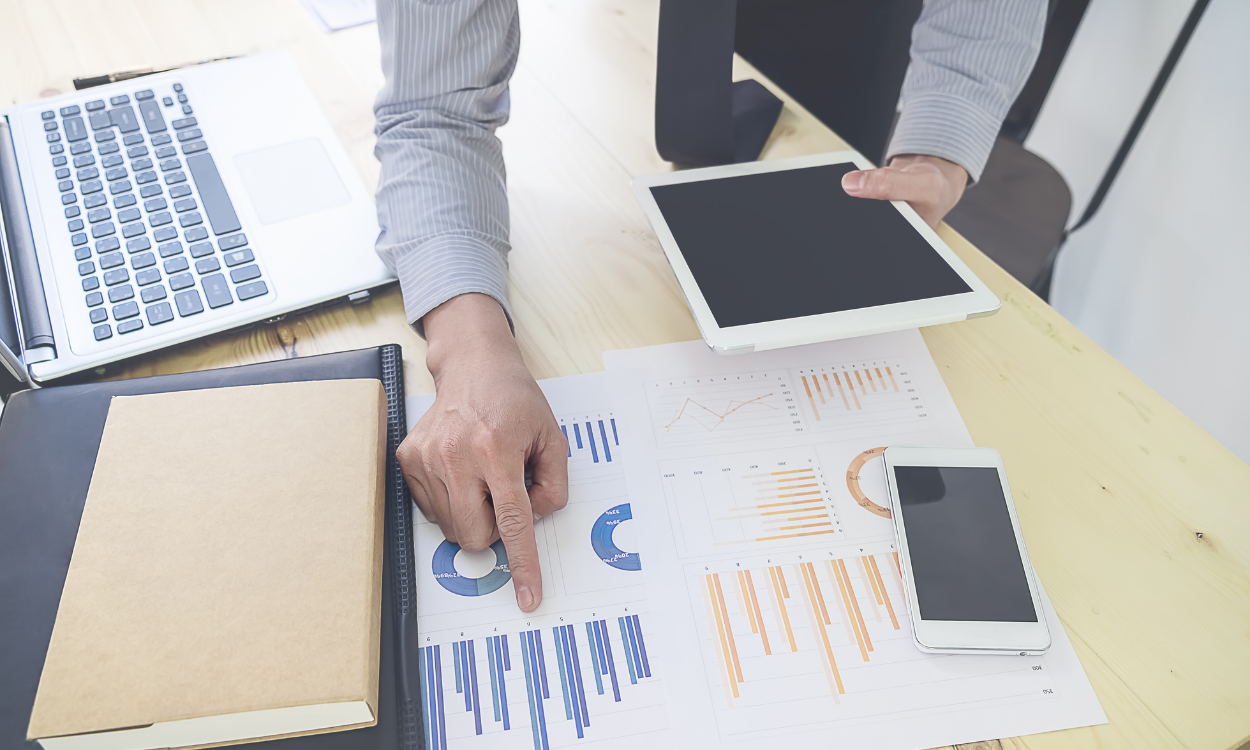 A person in a professional office setting points to data charts on a paper document while holding a tablet.