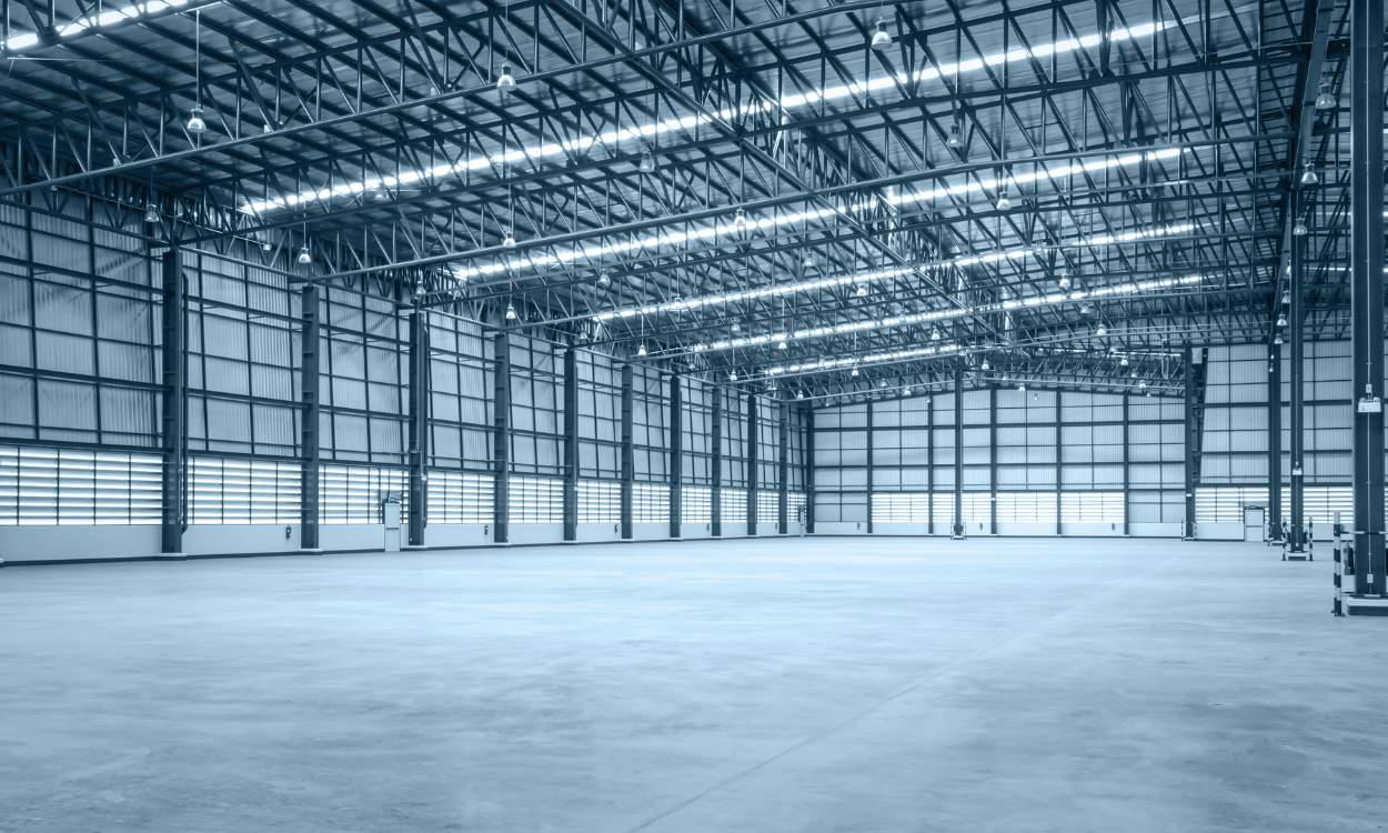 A large, empty industrial warehouse with a high steel-truss ceiling, concrete flooring, and bright overhead lighting.