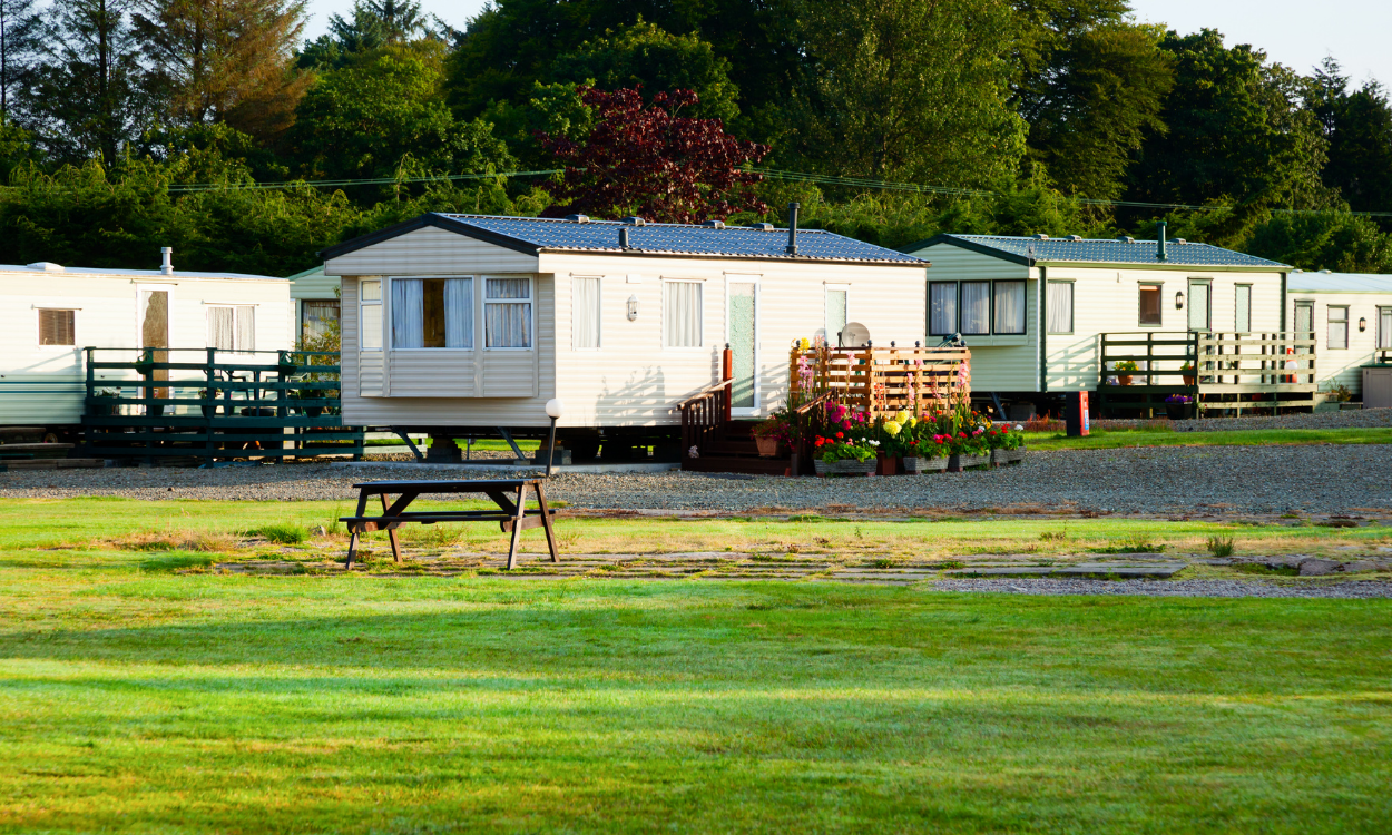 Mobile homes parked in a grassy area, with trees in the background. A picnic table sits in front of the homes.