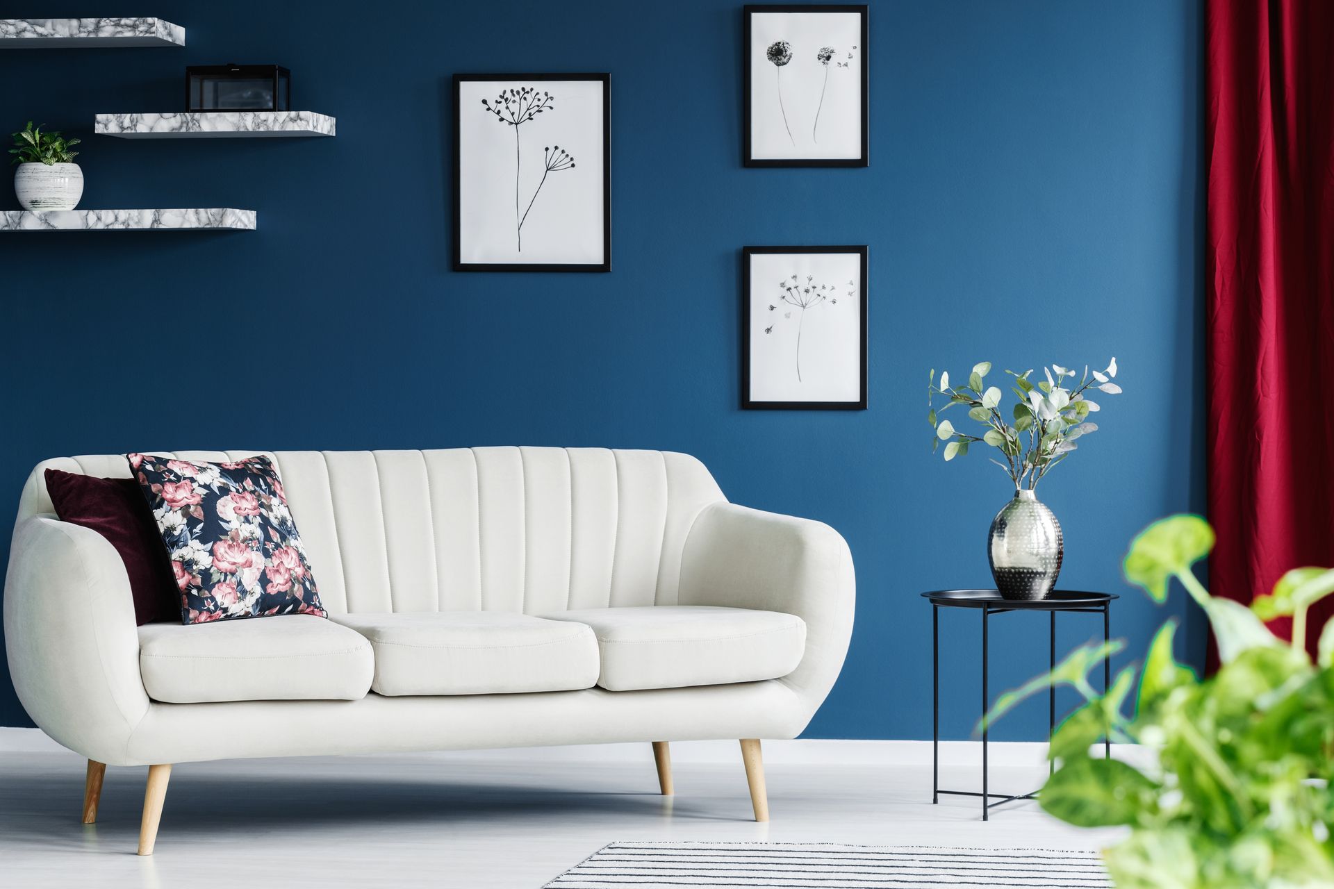 A white modern sofa sits in front of a deep blue wall decorated with three framed floral sketches and floating shelves.