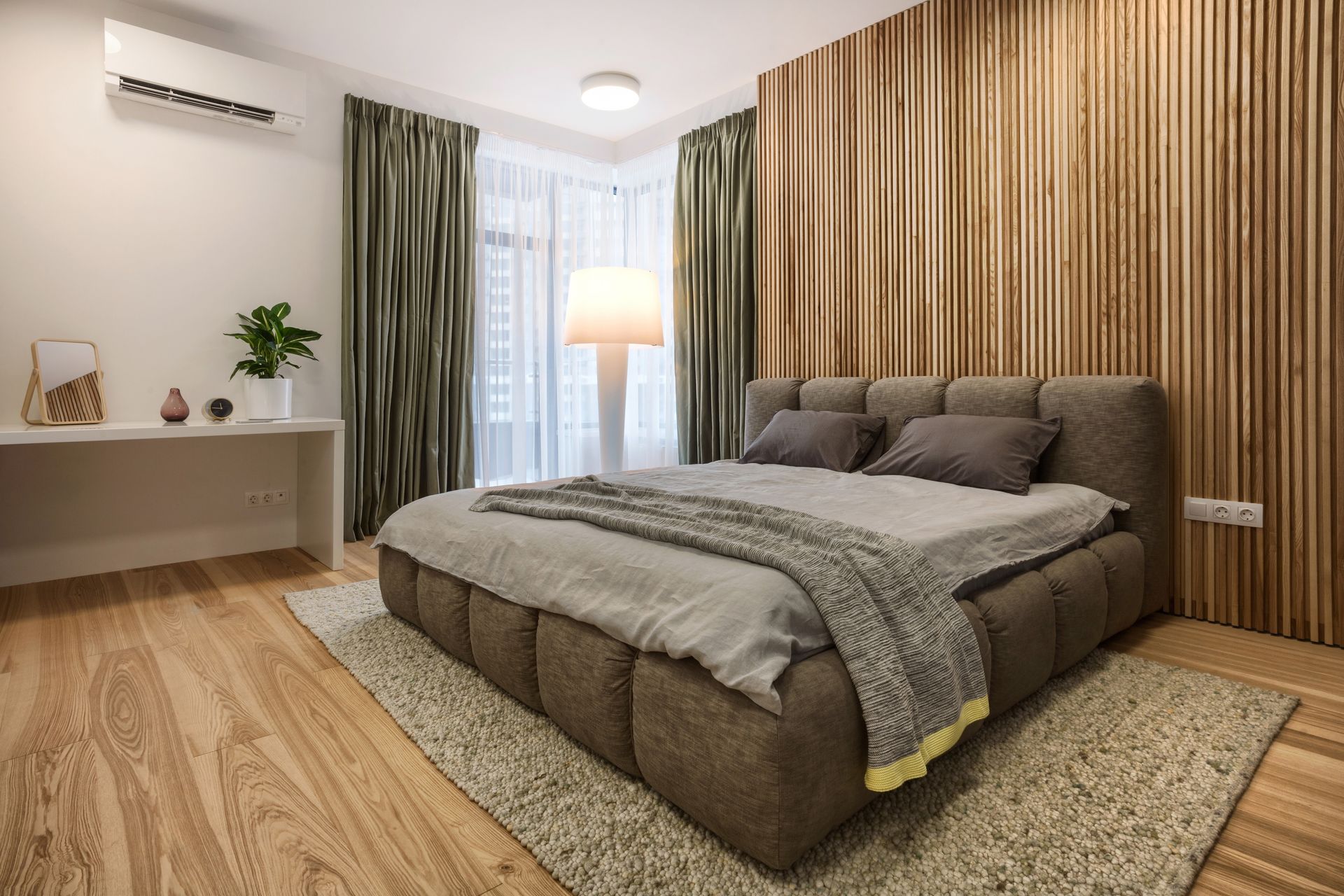 A modern bedroom with a brown upholstered bed, wooden slatted wall, green floor-length curtains, and a light-colored rug.