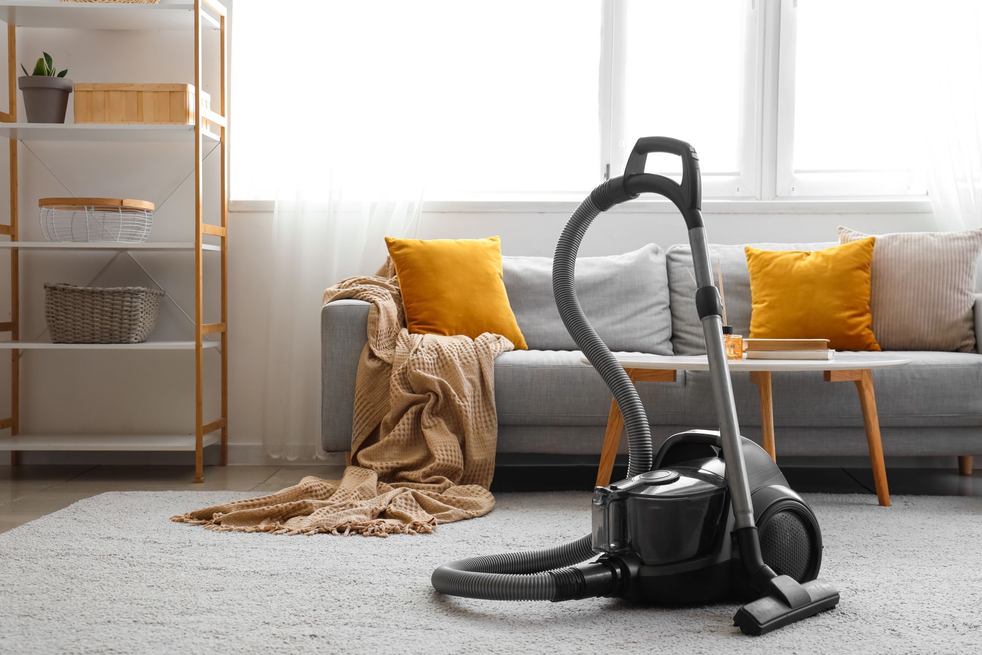 A black vacuum cleaner sits on a gray carpet in front of a sofa with yellow throw pillows and a tall shelving unit.