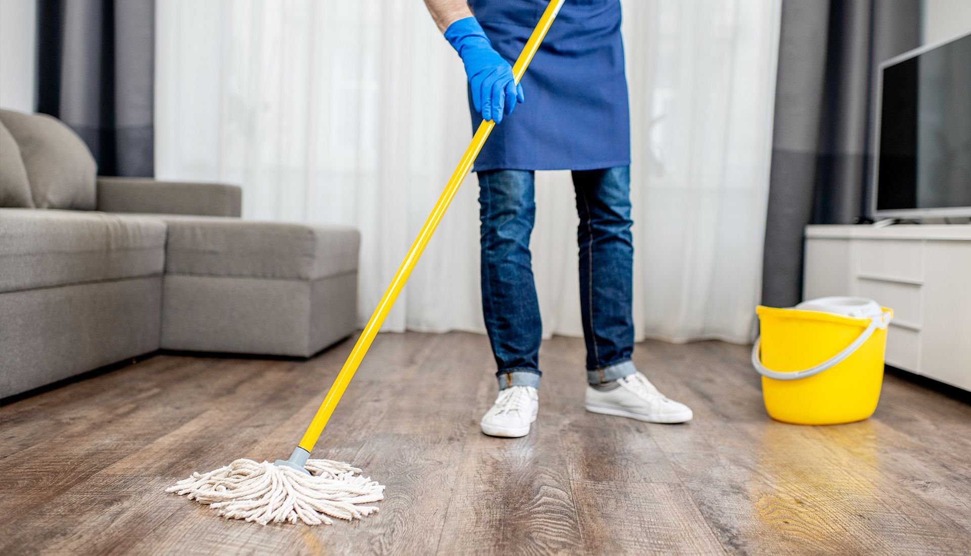 A person in blue jeans and an apron mopping a wooden floor next to a yellow bucket in a living room.
