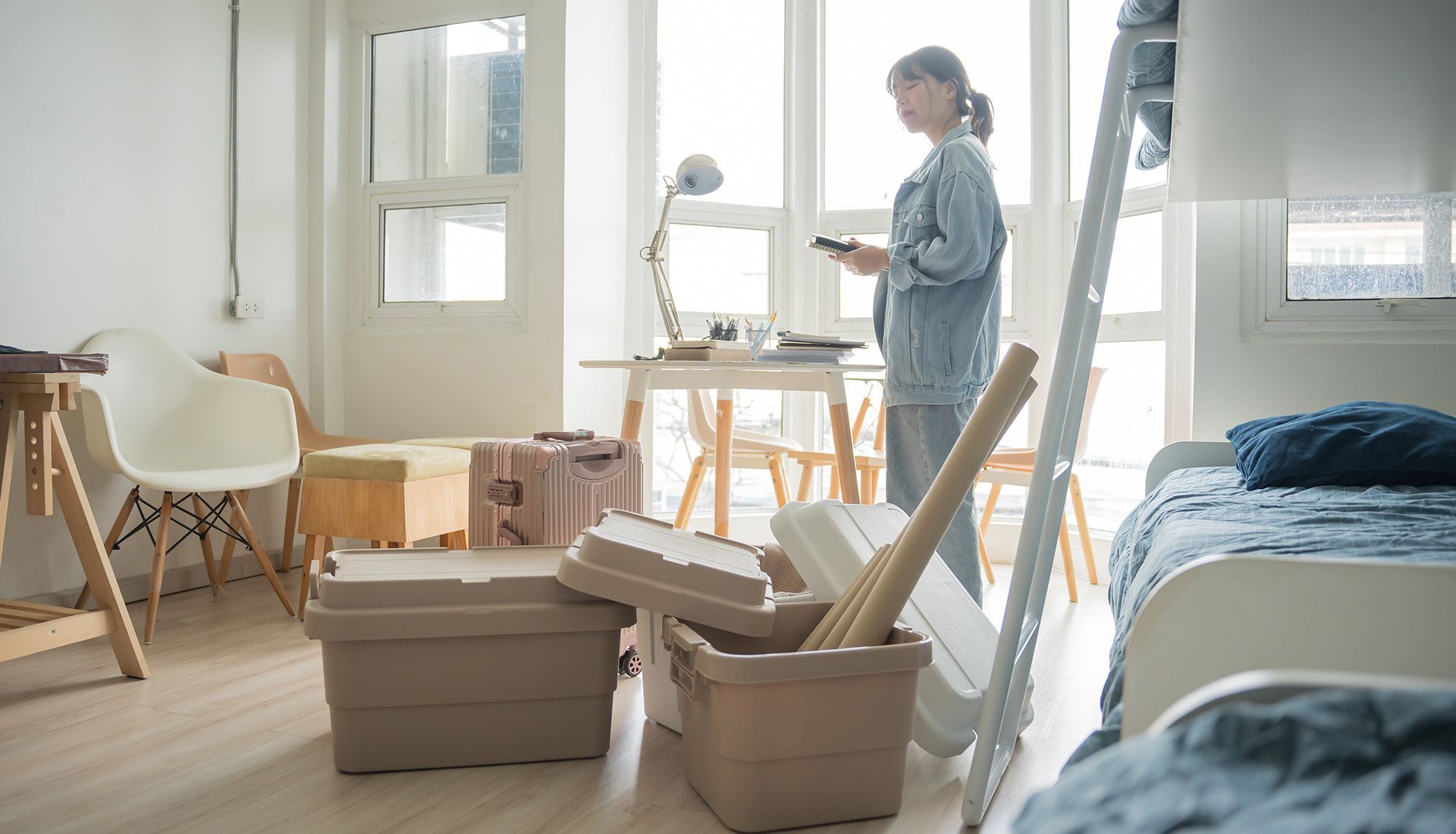 A person stands in a brightly lit, minimally furnished room filled with stacked plastic storage bins and moving supplies.