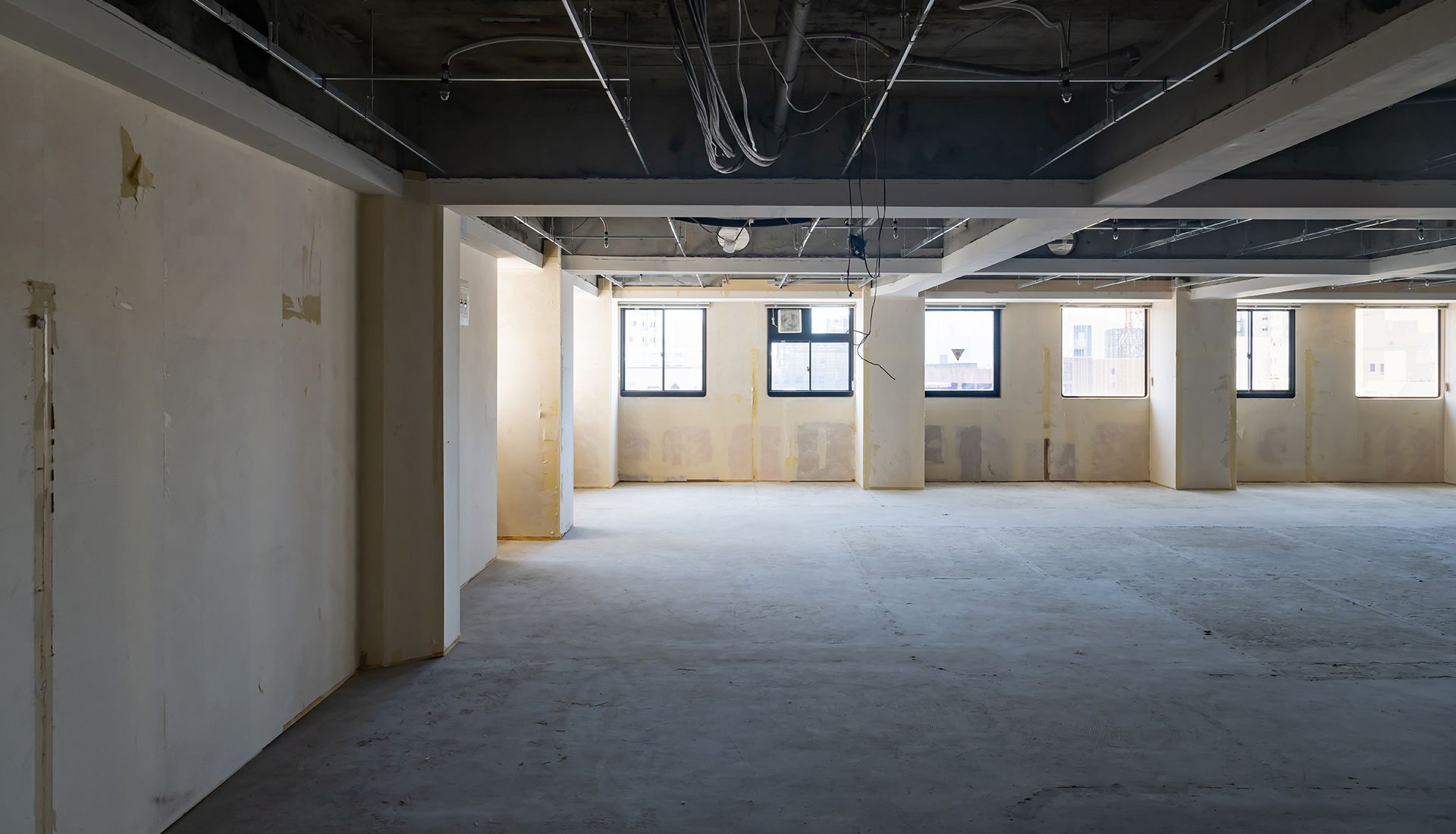 An open-concept, unfinished office space with concrete floors, white walls, support columns, and exposed ceiling wiring.