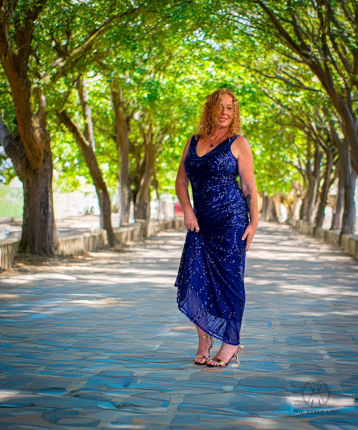 A woman in a blue dress is standing on a sidewalk surrounded by trees.