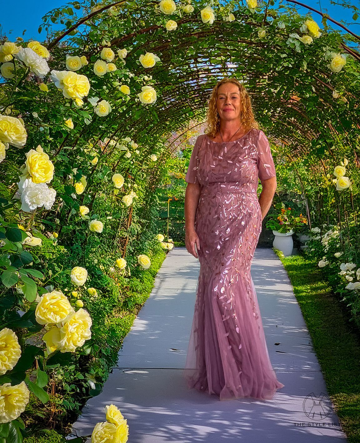 A woman in a pink dress is standing under an archway of yellow roses.