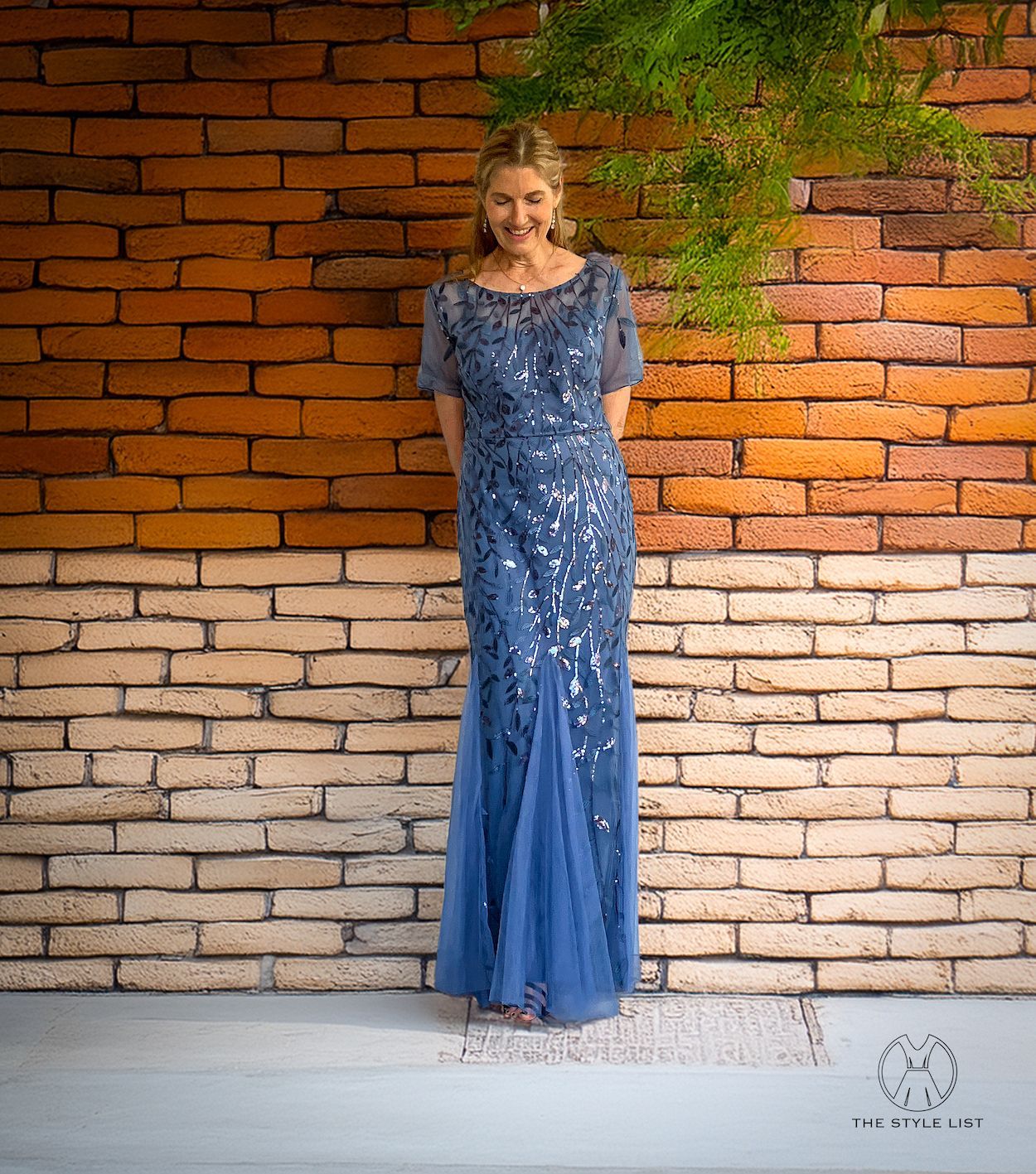 A woman in a blue dress is standing in front of a brick wall.