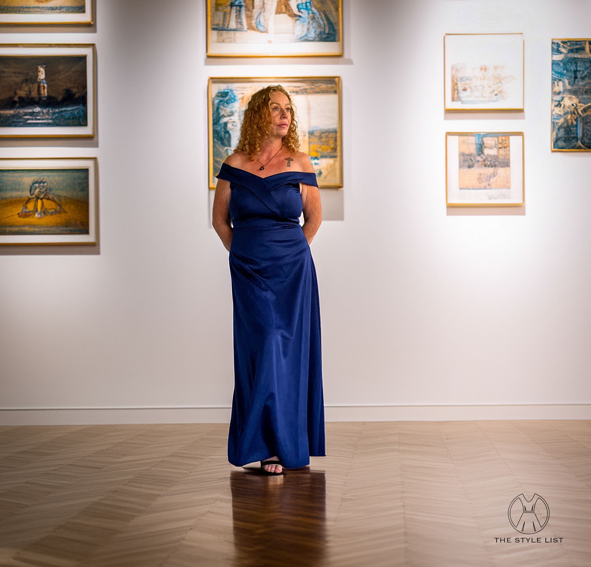 A woman in a blue dress stands in front of a wall of paintings