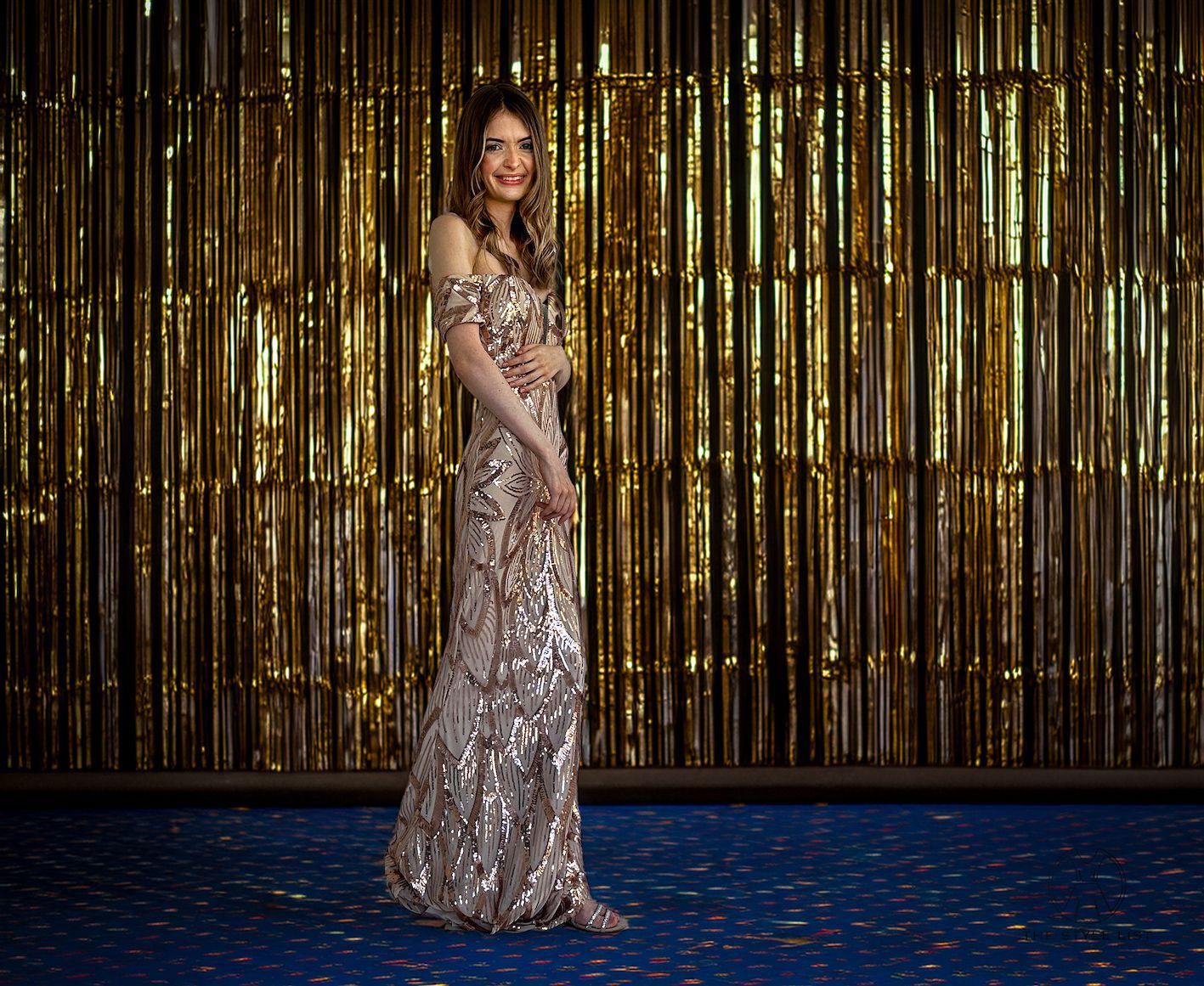 A woman in a long sequined dress is standing in front of a gold tinsel wall.