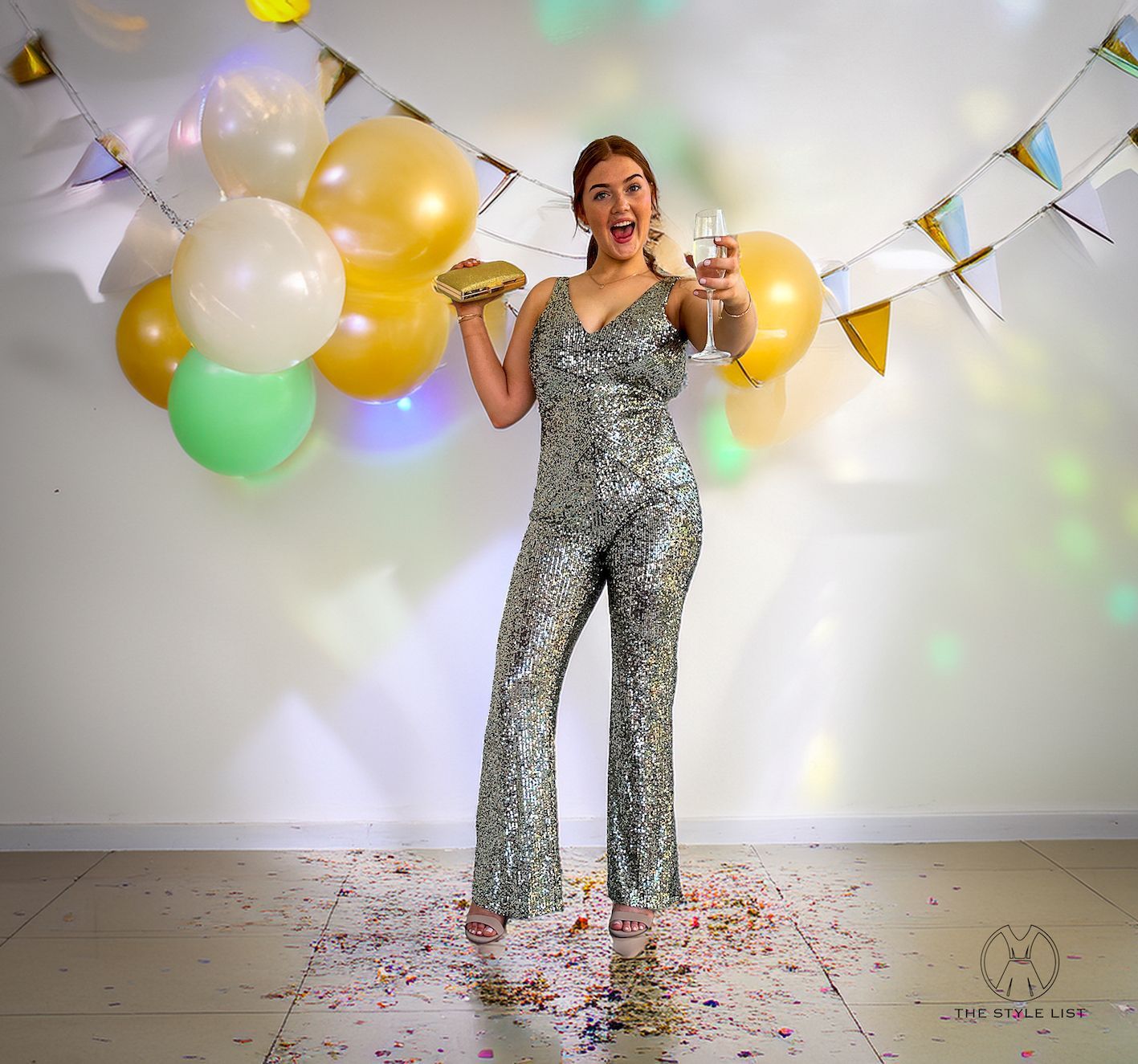 A woman in a sequined jumpsuit is holding balloons and a glass of champagne